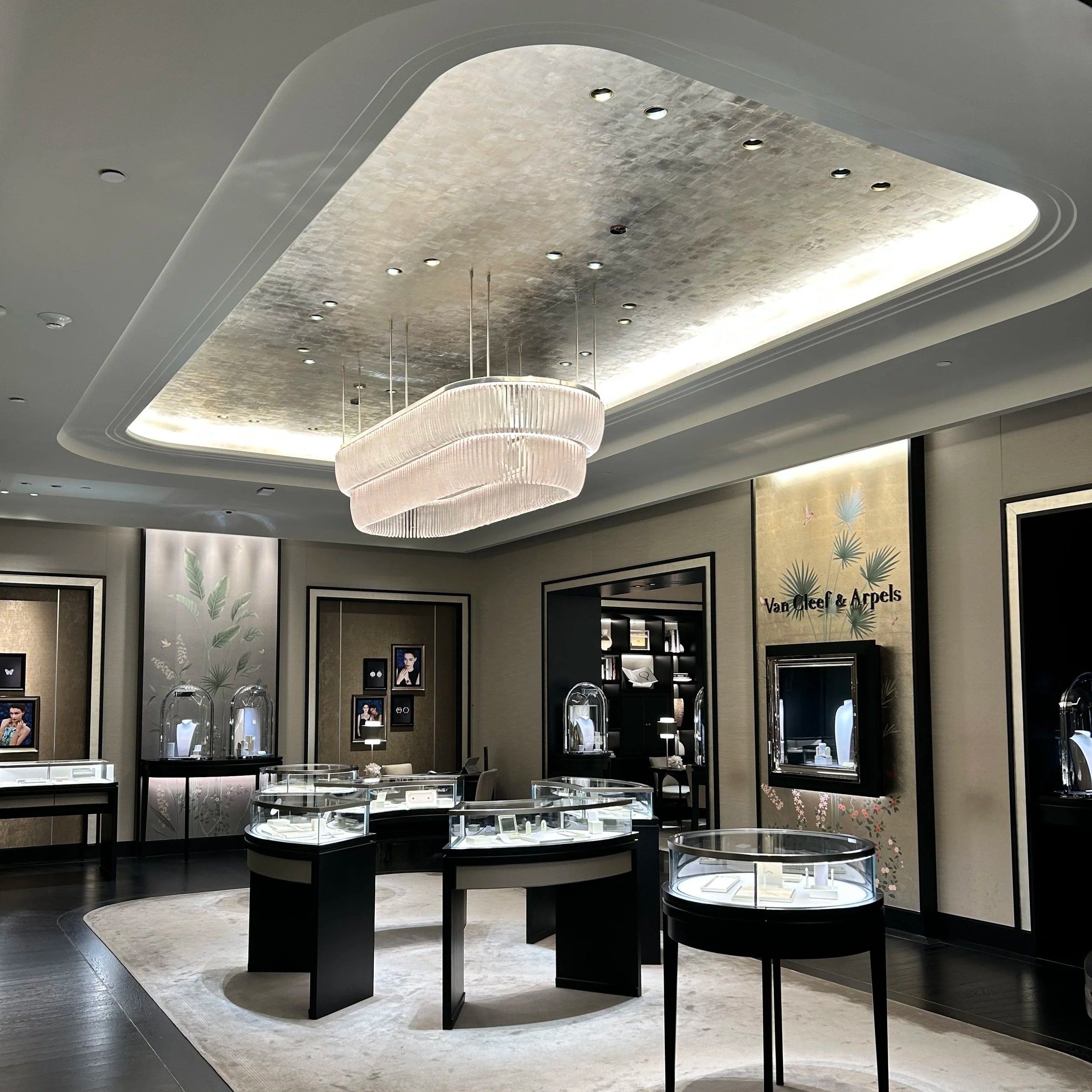 Luxury jewelry store display with glass cases and jewelry, decorative wall art featuring leaves, and a modern chandelier ceiling light.