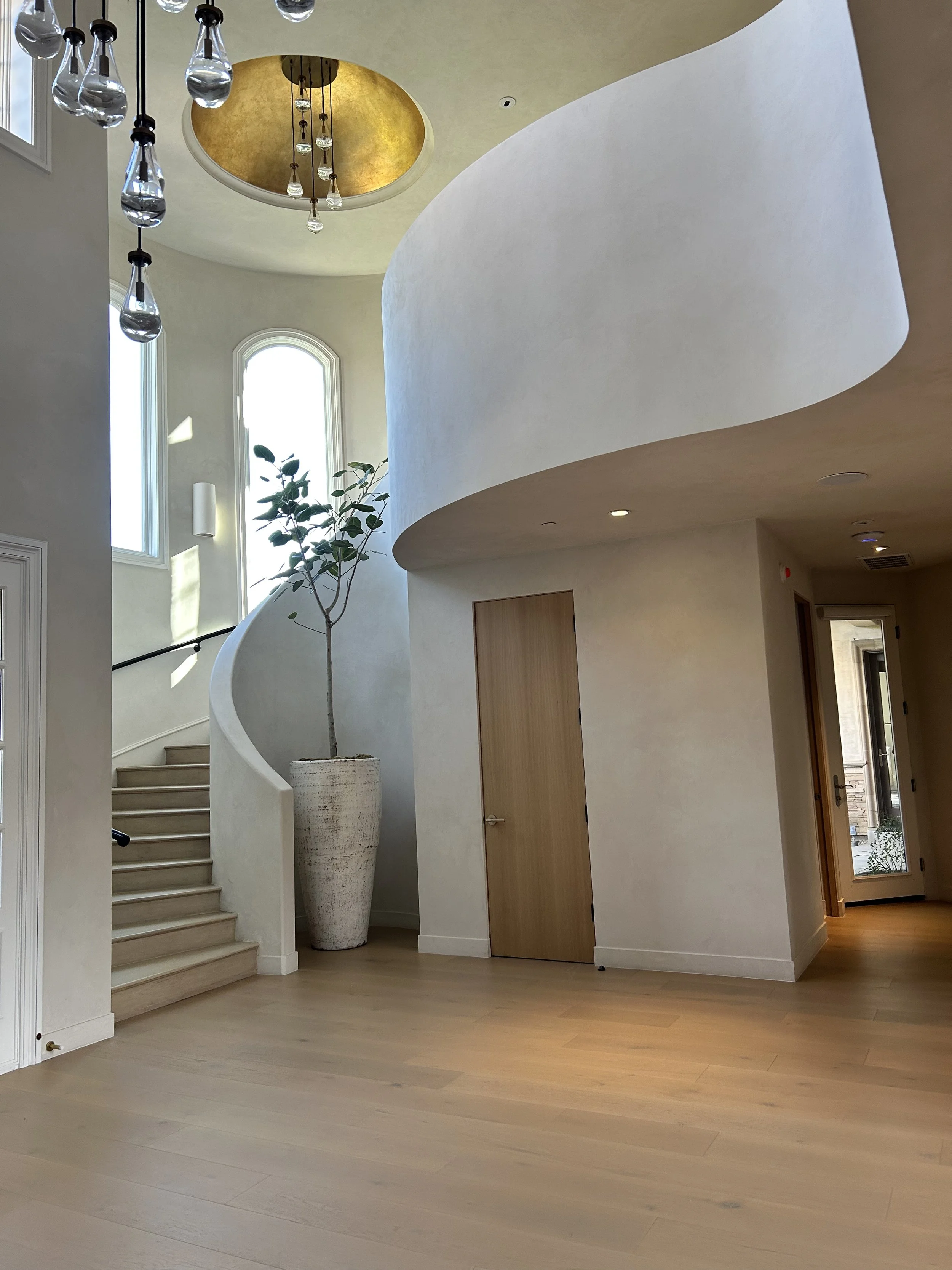Interior of a modern home with curved wall, wooden staircase, tall windows, and minimalist decor including a potted plant, chandelier, and pendant lights.