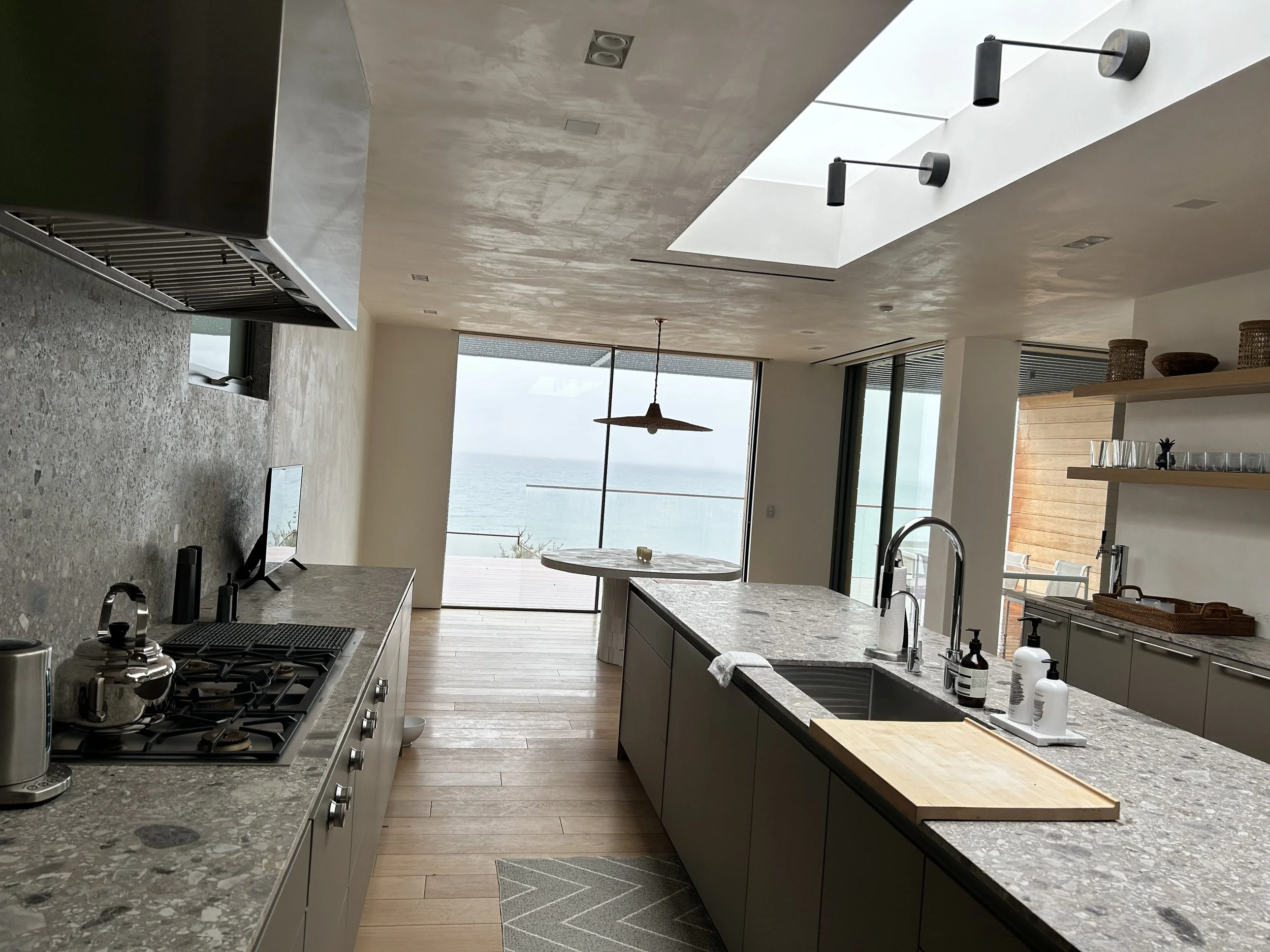 Modern kitchen with gray cabinets, marble countertops, a gas stove, and large windows overlooking the ocean. There is a dining table with a wooden top, a hanging pendant light, and decorative baskets on a wooden shelf. The ceiling has skylights and b