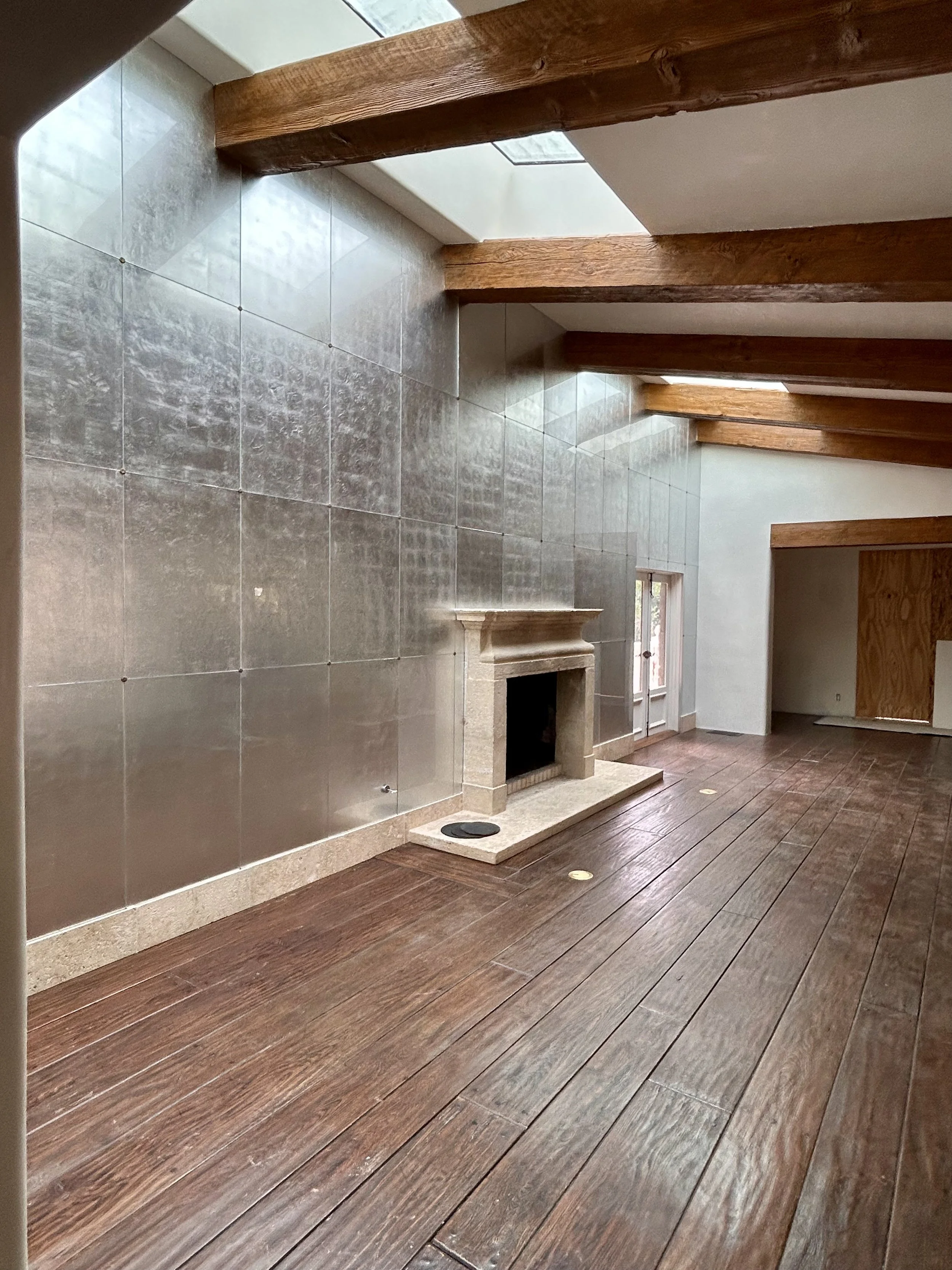 Interior of a room with wooden beams, tiled wall, and wood flooring, featuring a fireplace with a stone mantel and a paneled sliver leaf wall. 