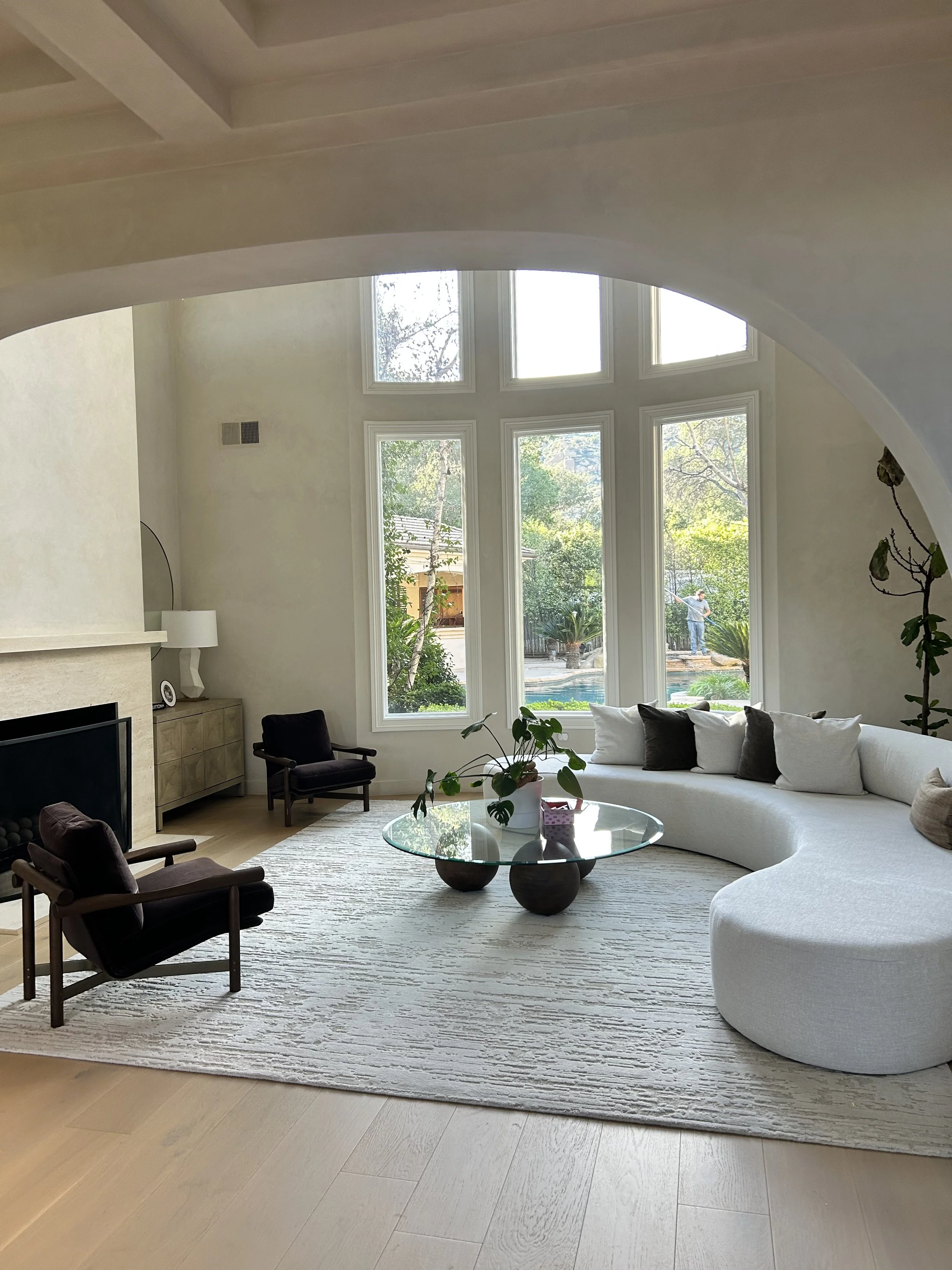 Living room with large stained glass window, white curved sofa with pillows, glass coffee table, two wooden armchairs, a white lamp, plants, and a fireplace with a TV on top.