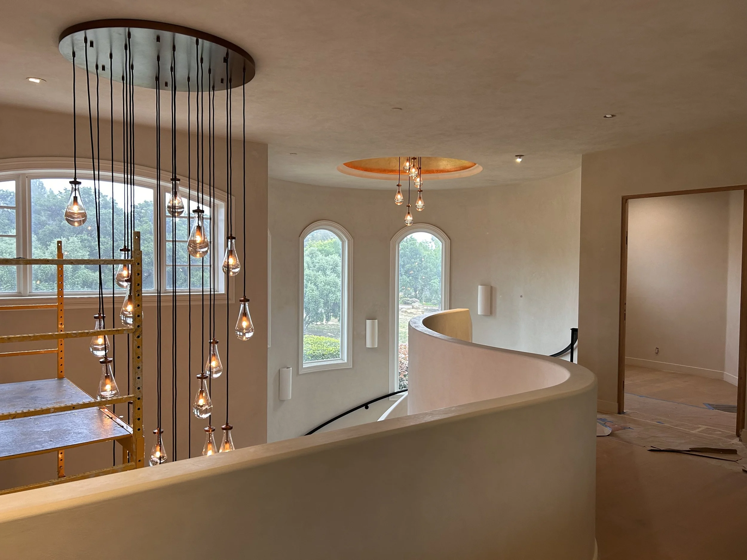 Interior of a house under construction featuring large arched windows, modern ceiling lights, a curved staircase, and a decorative ceiling feature with hanging pendant lights.