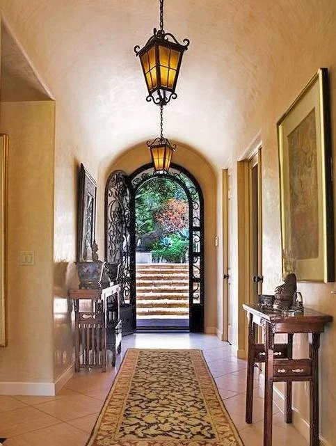 Interior hallway with tiled floor, a decorative runner rug, and a wrought iron arched door leading to stairs outside. Two hanging lantern-style lights, side tables with decorative items, and framed art on the walls.