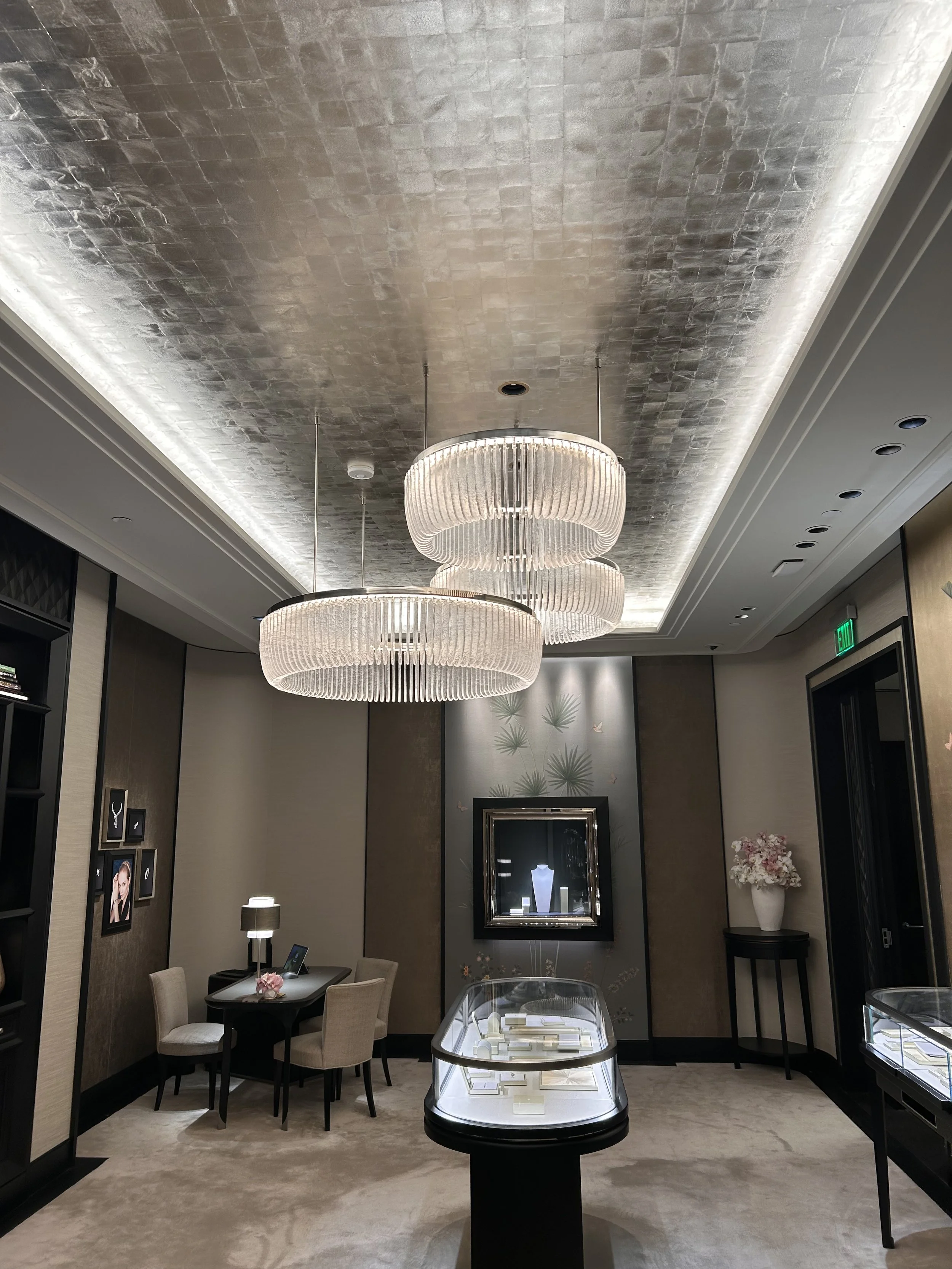Interior of a jewelry store with display cases showcasing jewelry, a seating area with chairs, decorative wall art, and a large chandelier lighting fixture hanging from a textured metallic ceiling.