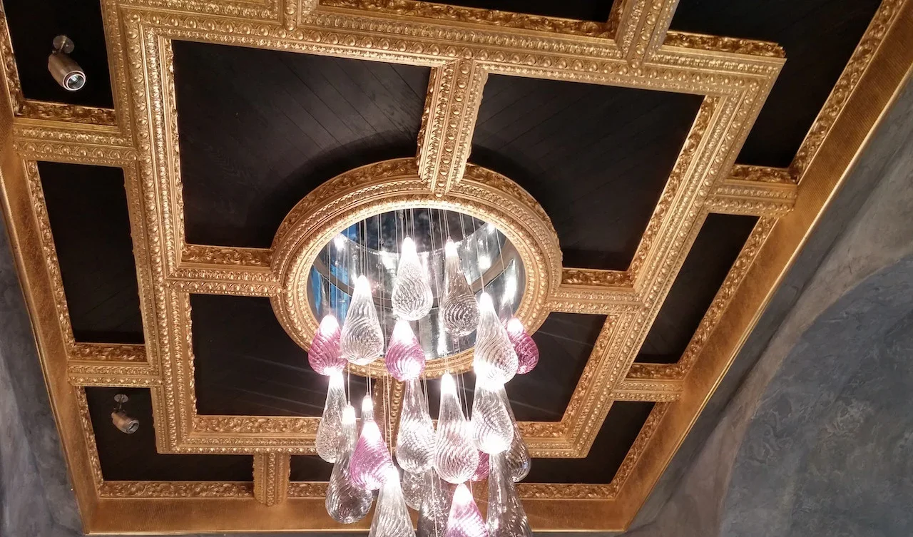 A luxurious ceiling with an ornate gold-trimmed black pattern around a central mirror and an elaborate chandelier with glass teardrop-shaped ornaments hanging from it.