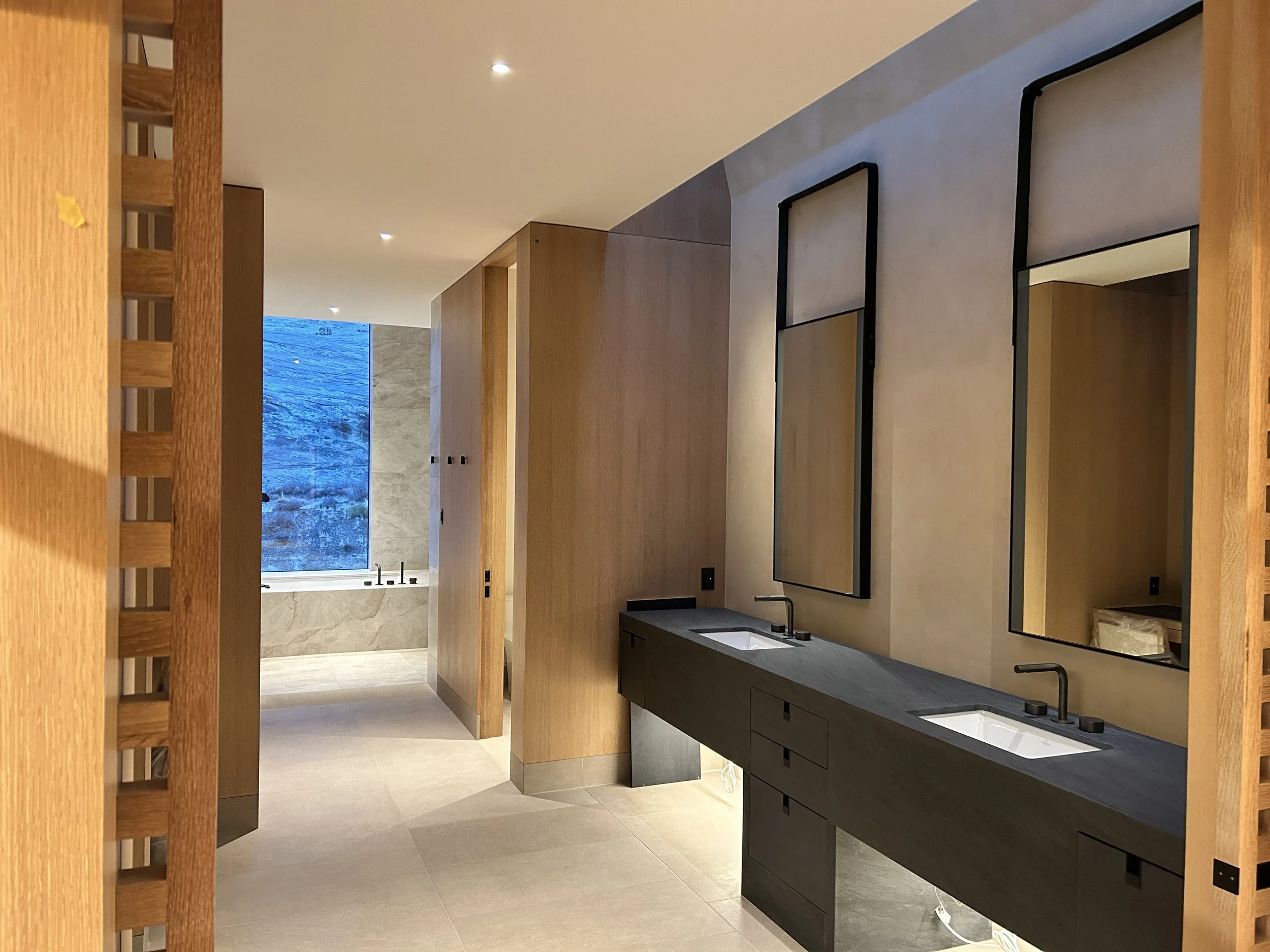 Modern bathroom with black double sinks, large rectangular mirrors, wooden cabinets, beige tile flooring and walls, and a window showing a mountain landscape.