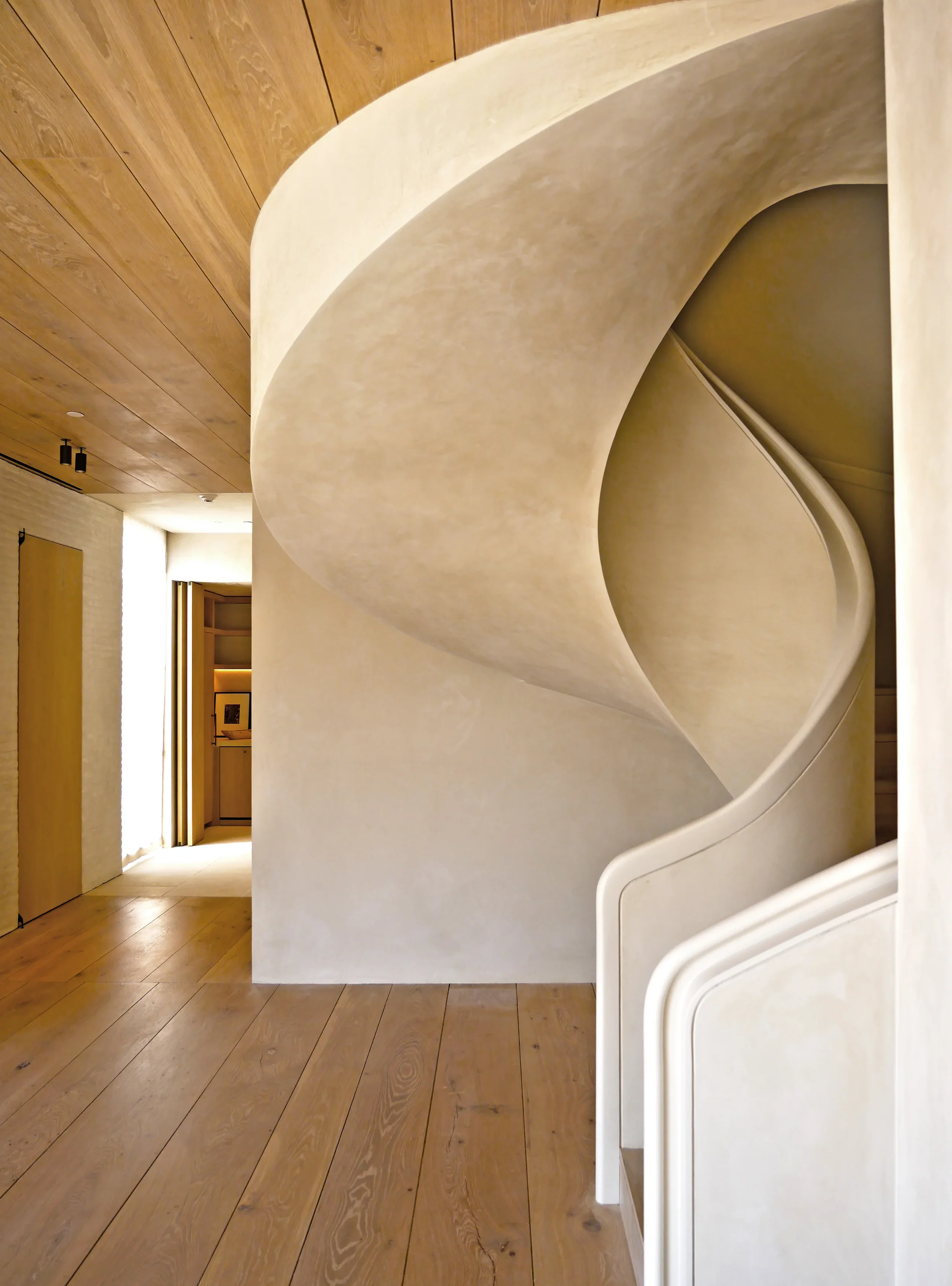 A modern interior space featuring a curved beige wall, wooden ceiling, wooden flooring, and a doorway with a warm, inviting glow.
