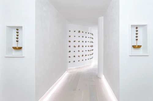 Minimalist white hallway with two wall niches holding stacked decorative objects, and a wall with small sculptures arranged in a grid pattern.