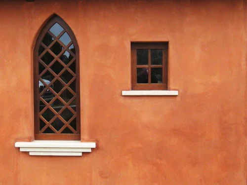 Close-up of an orange stucco wall with two windows: a tall, arched window with a crisscross wooden lattice and a small, rectangular window, both with white sills.