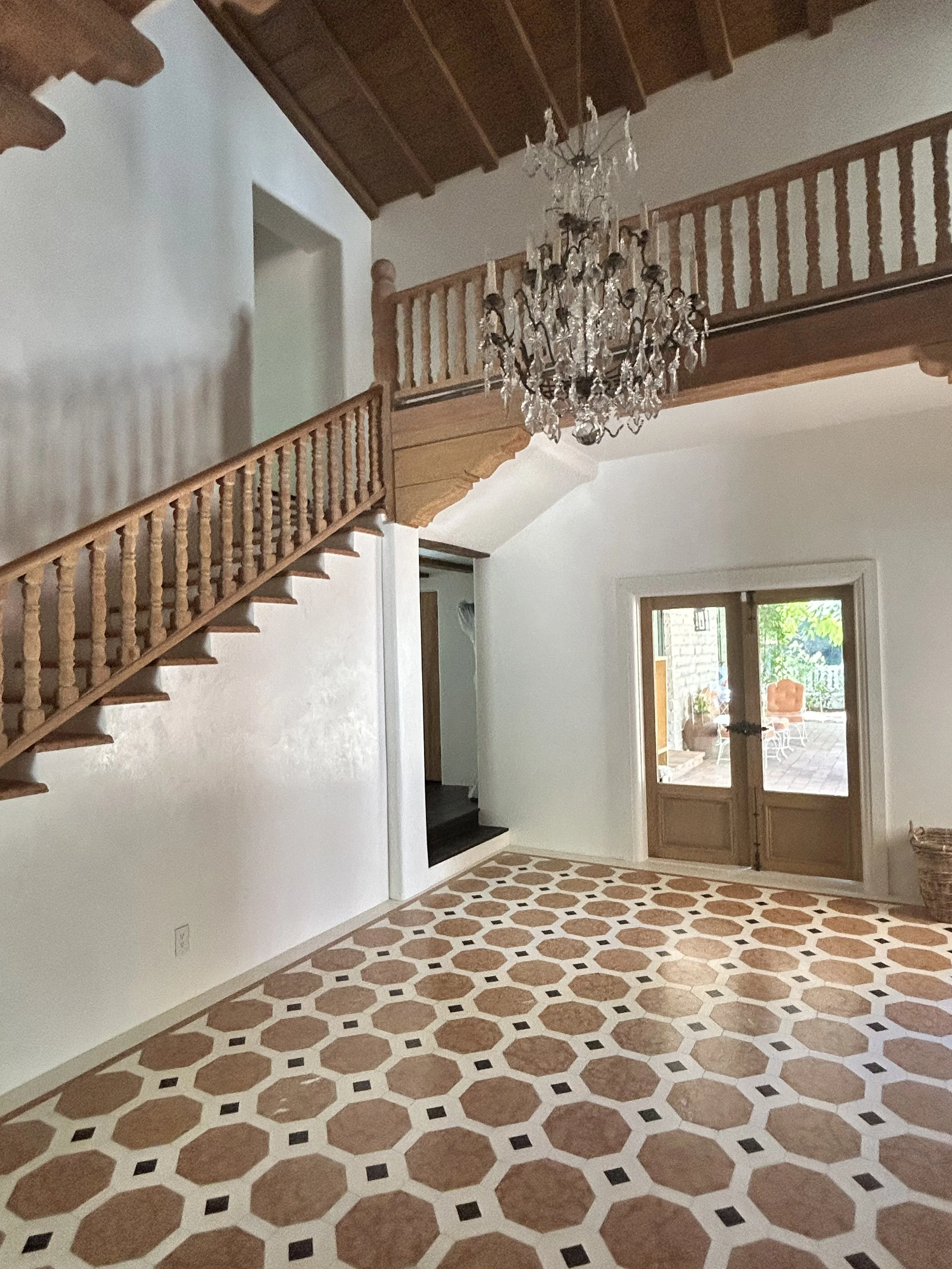 Interior view of a room with patterned tile flooring, wooden stairs leading to a second floor, a chandelier hanging from the ceiling, and double glass doors opening to an outdoor area.