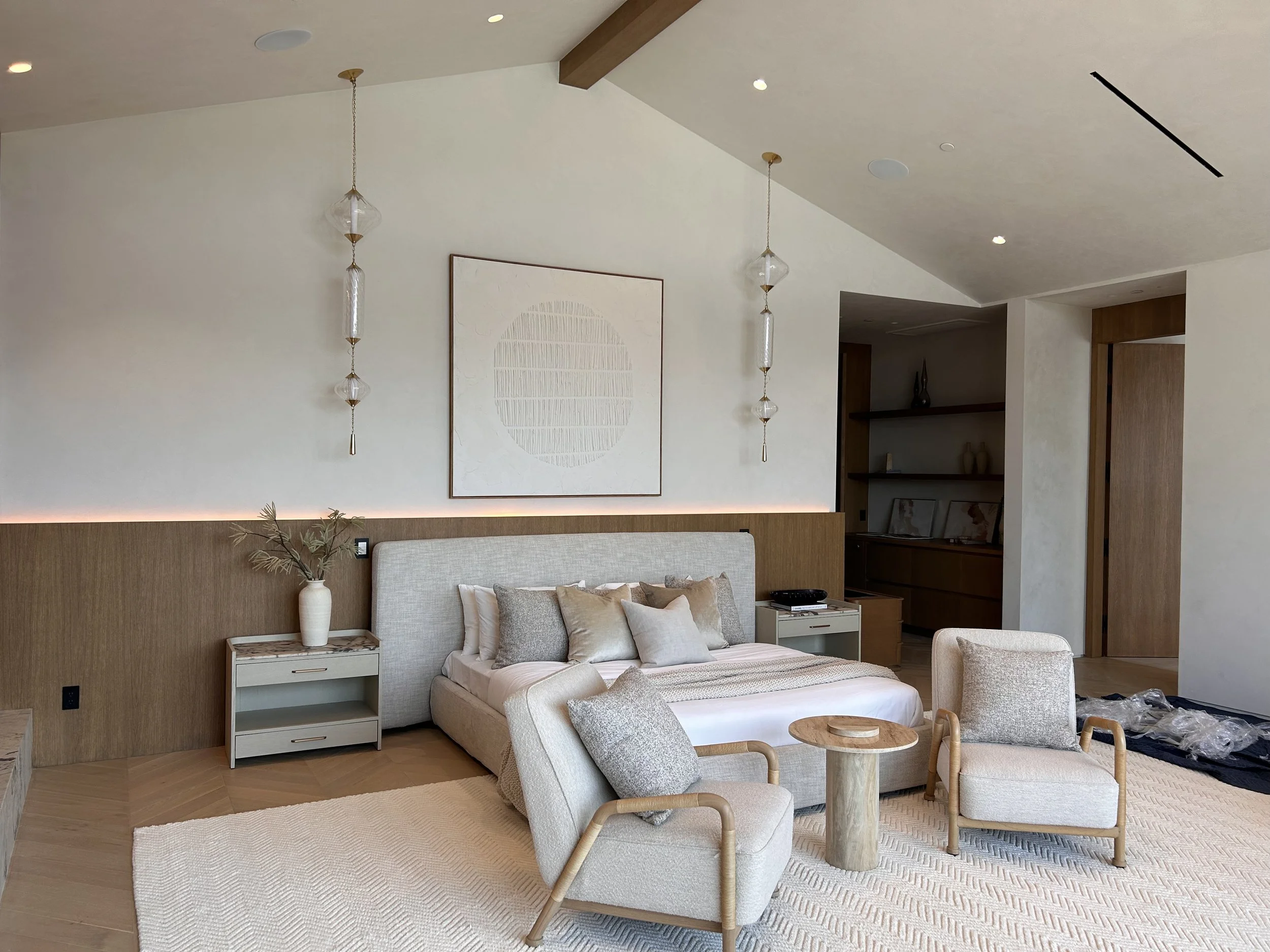 A modern living room with a beige sofa, a white side table, and a light-colored armchair. There are decorative pillows on the sofa, a potted plant on the side table, and a round wooden coffee table. The wall behind has minimalist art and hanging pend
