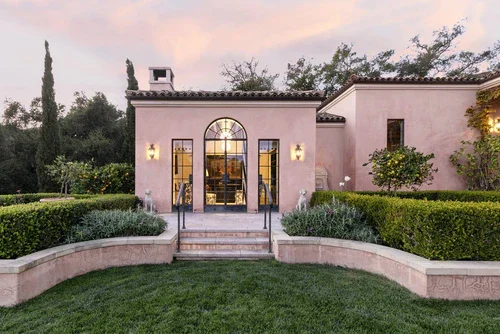 Front view of a pink stucco house with an arched glass door and black window frames, surrounded by manicured lawns and garden bushes at sunset.