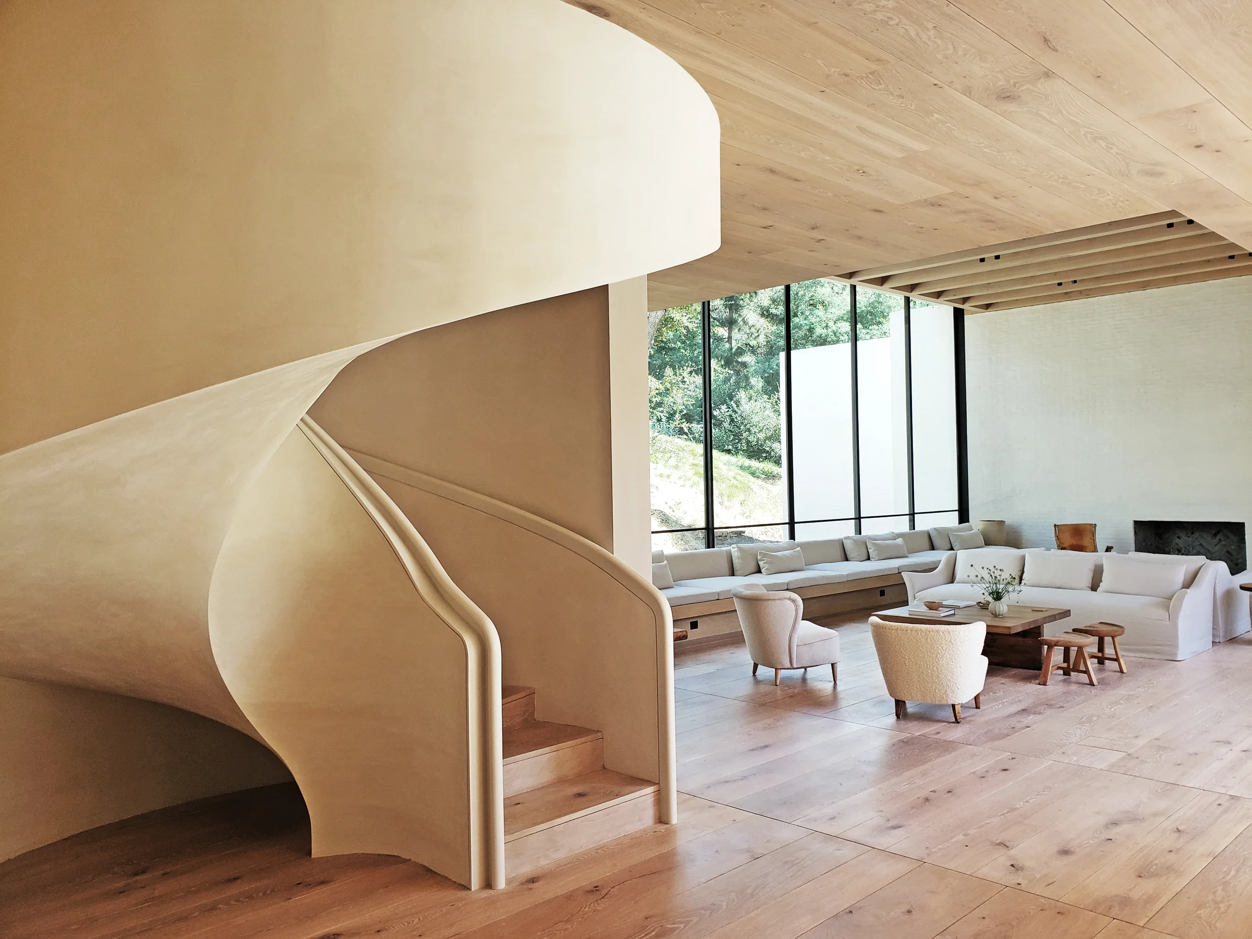 Modern living room with beige walls, large glass windows showing trees outside, and a light wood ceiling. Features a white L-shaped sofa with pillows, a wooden coffee table, and two armchairs.