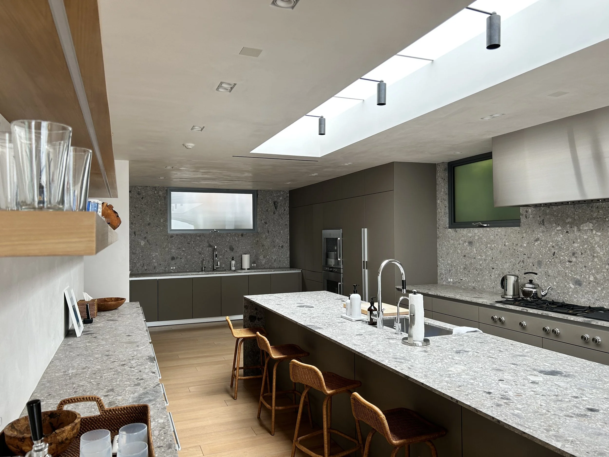 Modern kitchen with a large central island, grey cabinetry, granite countertops, a sink, and a stove. A window and skylight allow natural light to fill the space. There are bar stools along the island and hanging cylindrical pendant lights.