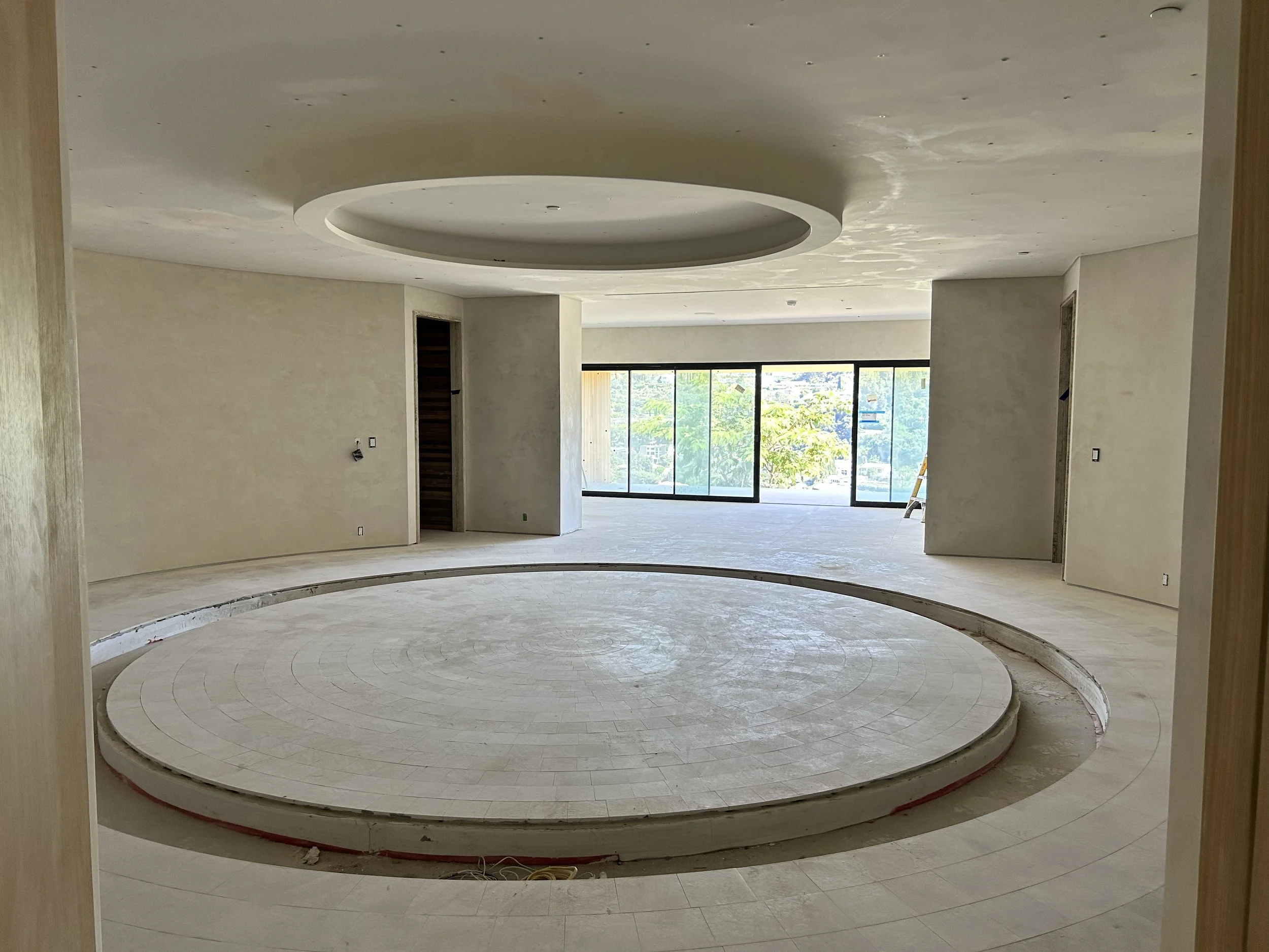Interior of a building under construction with a circular platform in the center, unfinished walls, large windows with a view of trees, and construction materials