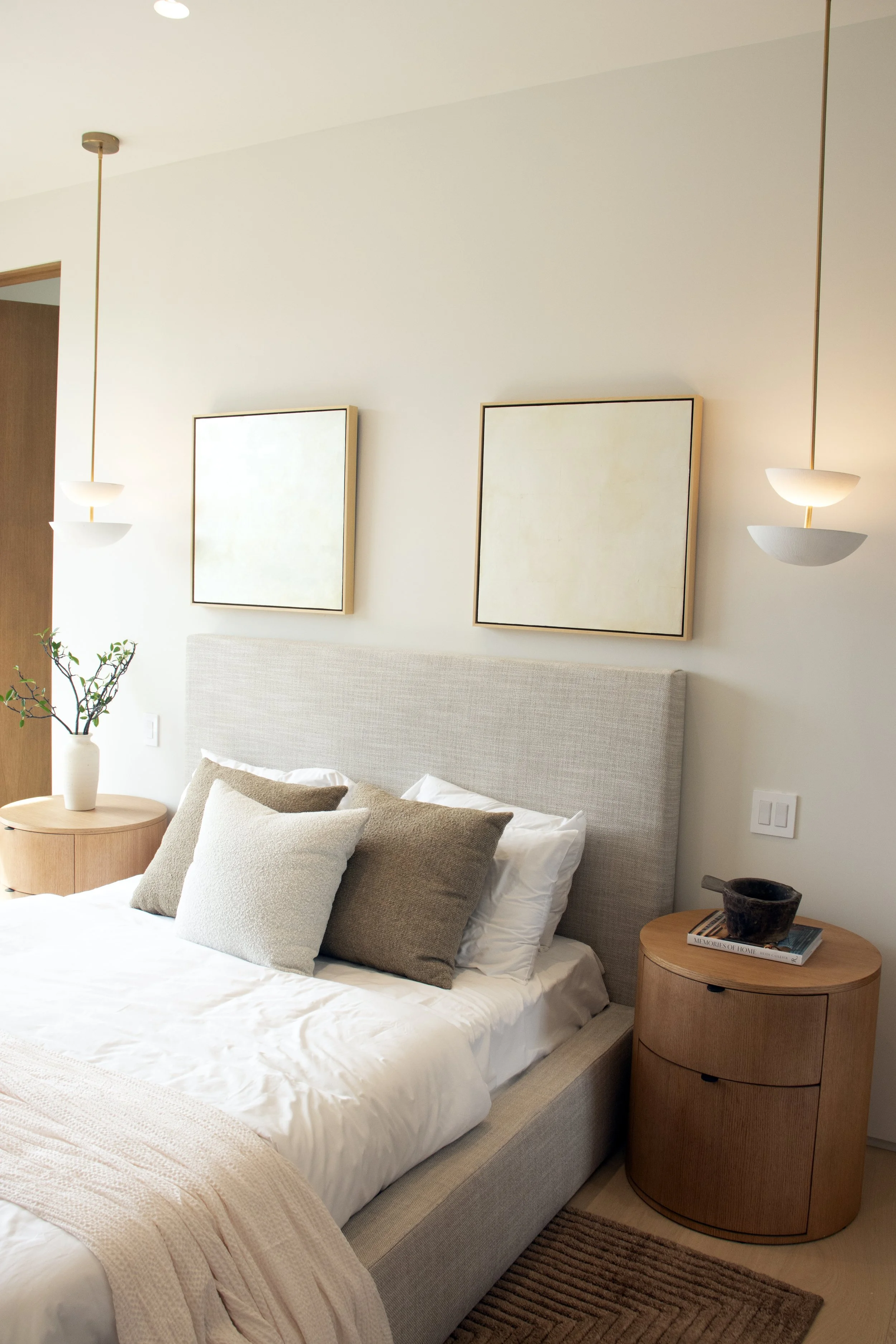 A neatly made bed with a beige upholstered headboard, white bedding, and several neutral-colored pillows. Flanking the bed are matching round wooden side tables; the right one has a decorative bowl and books. Two minimalist wall sconces with a modern design and soft lighting hang above the beds, and two framed artworks with simple borders are on the plain white wall behind.