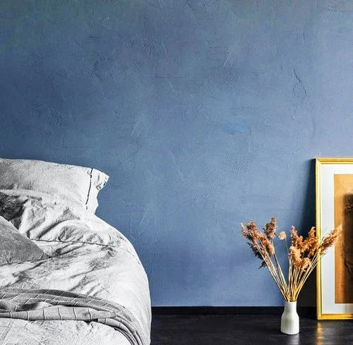 A bedroom with a blue textured wall, a bed with white and gray bedding, a white vase with dried flowers, and a gold-framed picture leaning against the wall.
