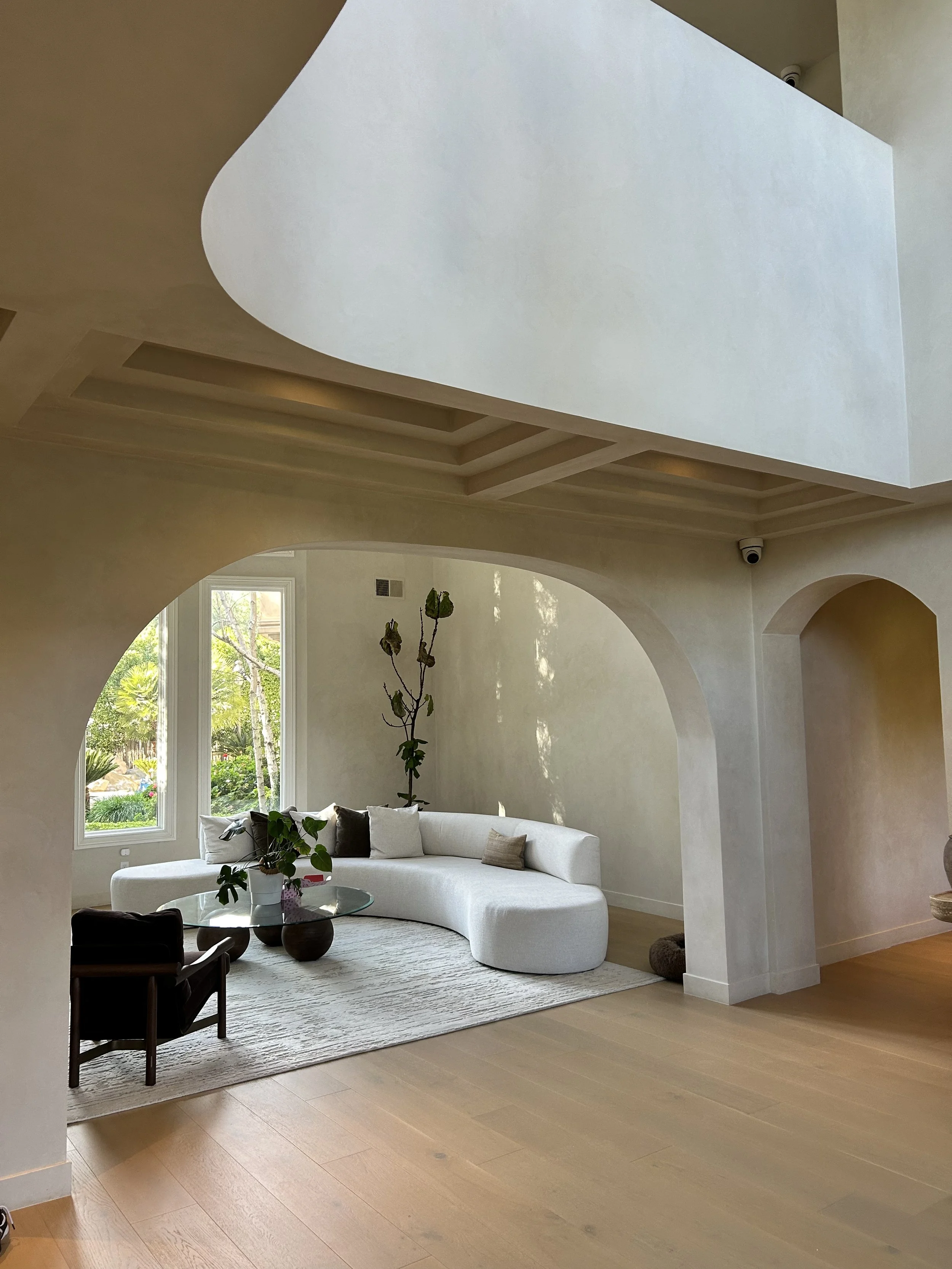Living room with large white curved sofa, glass coffee table, potted plant, and large windows showing outdoor greenery.