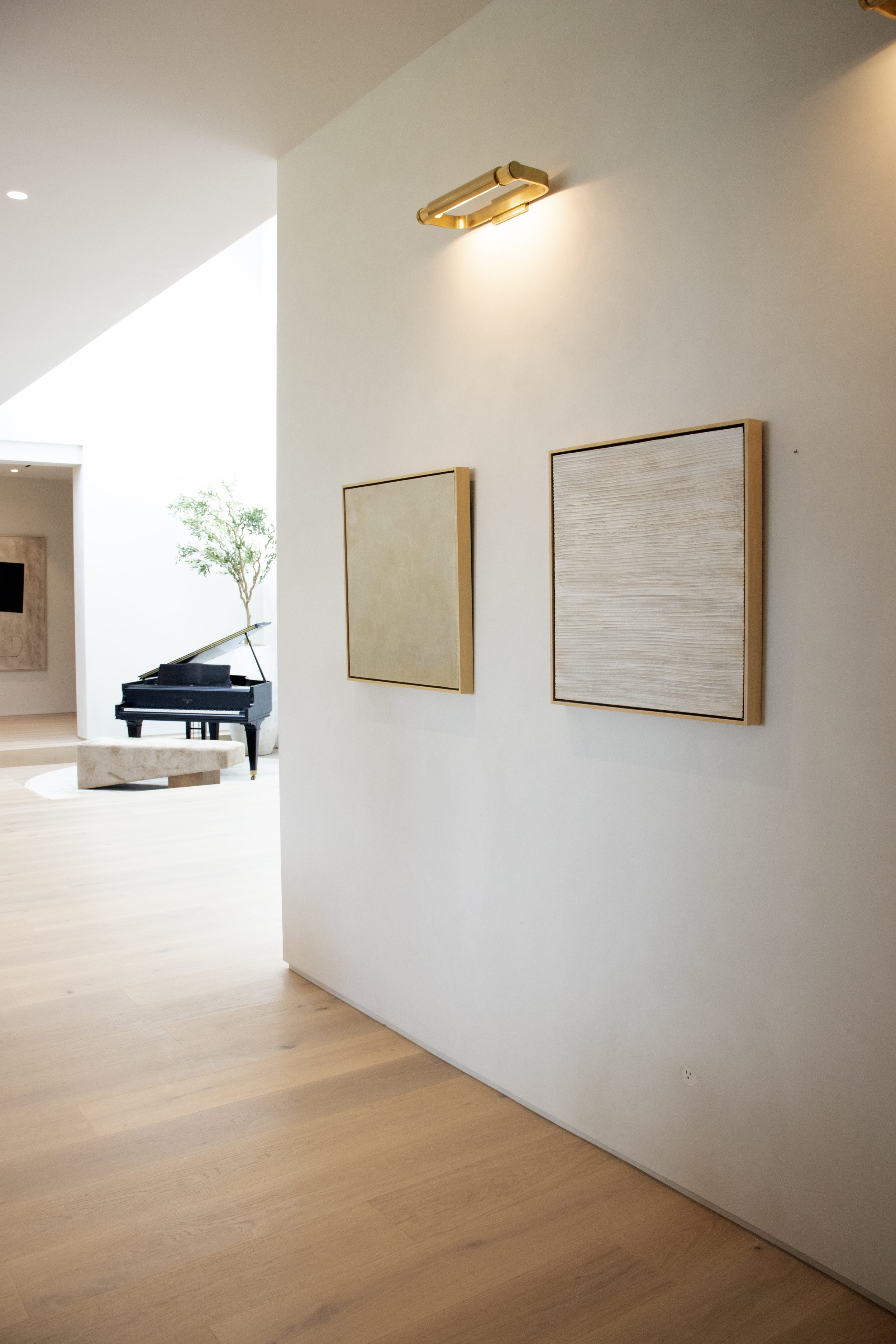 A modern interior with light wooden flooring, two framed abstract artworks on a white wall, and a black grand piano with a bench in a spacious room illuminated by natural light.