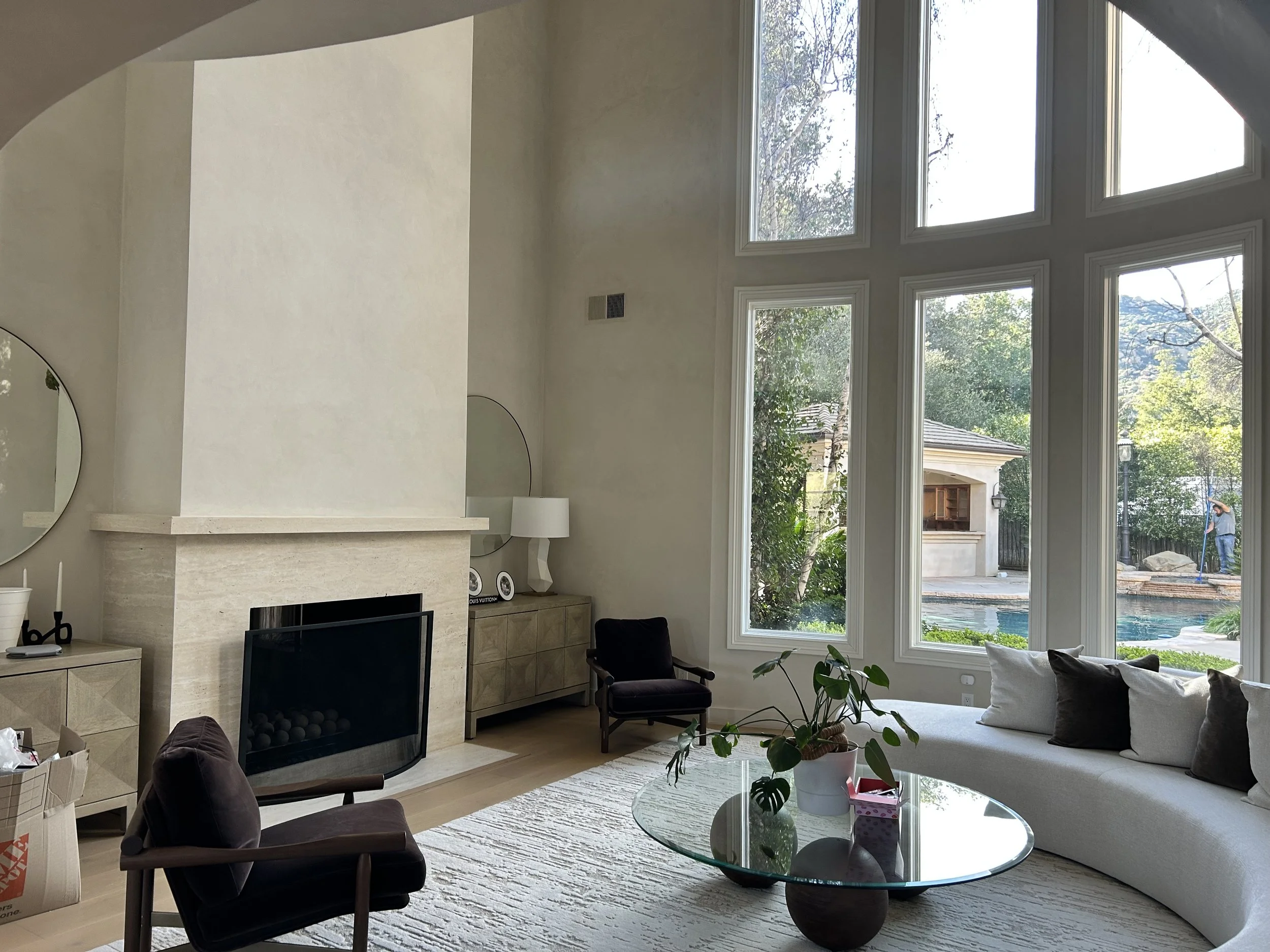 Living room with large windows showing a backyard with trees and mountains, a white sofa with pillows, a glass coffee table with plants, and chairs.