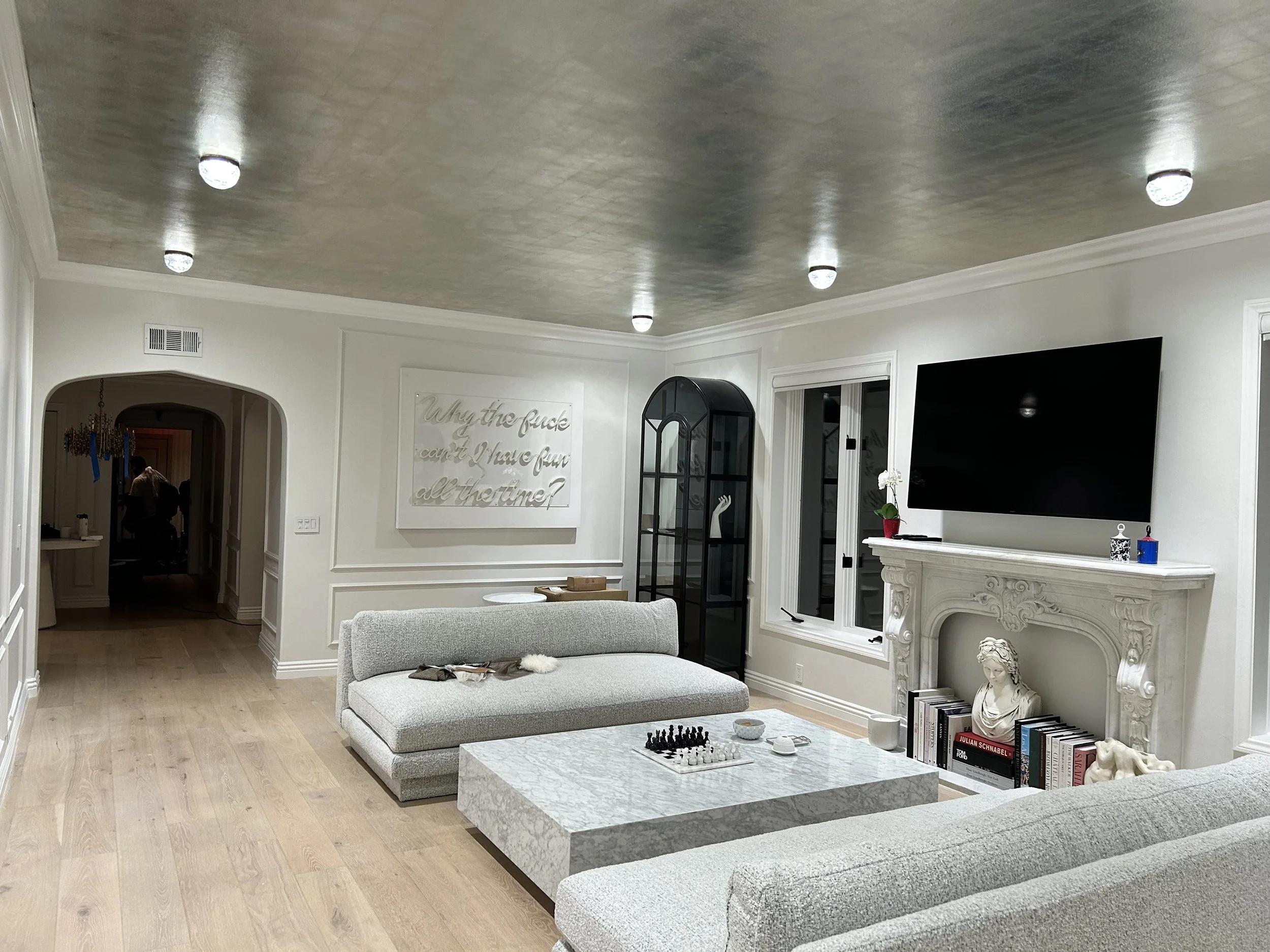 Living room with white walls and crown molding, featuring a gray sectional sofa and a marble coffee table with chess pieces. Decor includes books on shelves, a white bust, and a wall-mounted television, with a sign that says 'Why the fuck can't I hav
