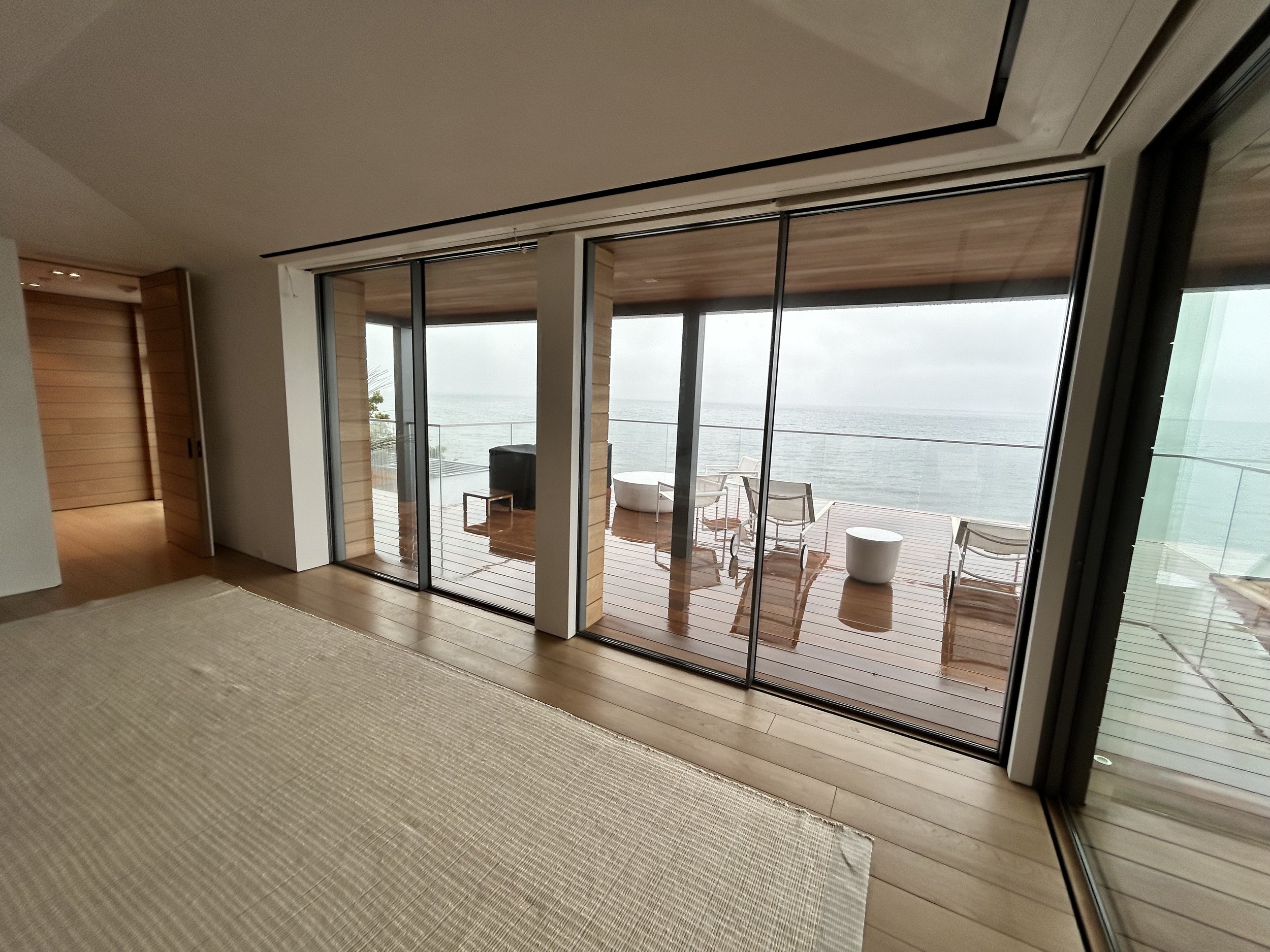 Interior view of a room with large sliding glass doors opening to a balcony overlooking the ocean, with outdoor furniture visible.