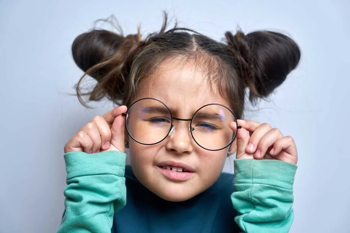 Young girl with messy hair styled in buns, holding glasses up to her face, squinting, wearing a blue shirt and green long-sleeve shirt
