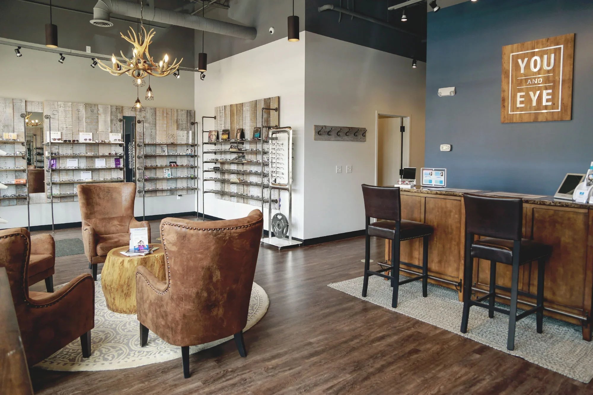 Interior of an optical store or eye clinic with display shelves of glasses, a reception desk, and seating area with brown velvet armchairs and a round wooden table. A decorative chandelier hangs from the ceiling, and there is a large sign on the blue wall that reads 'YOU AND EYE'.