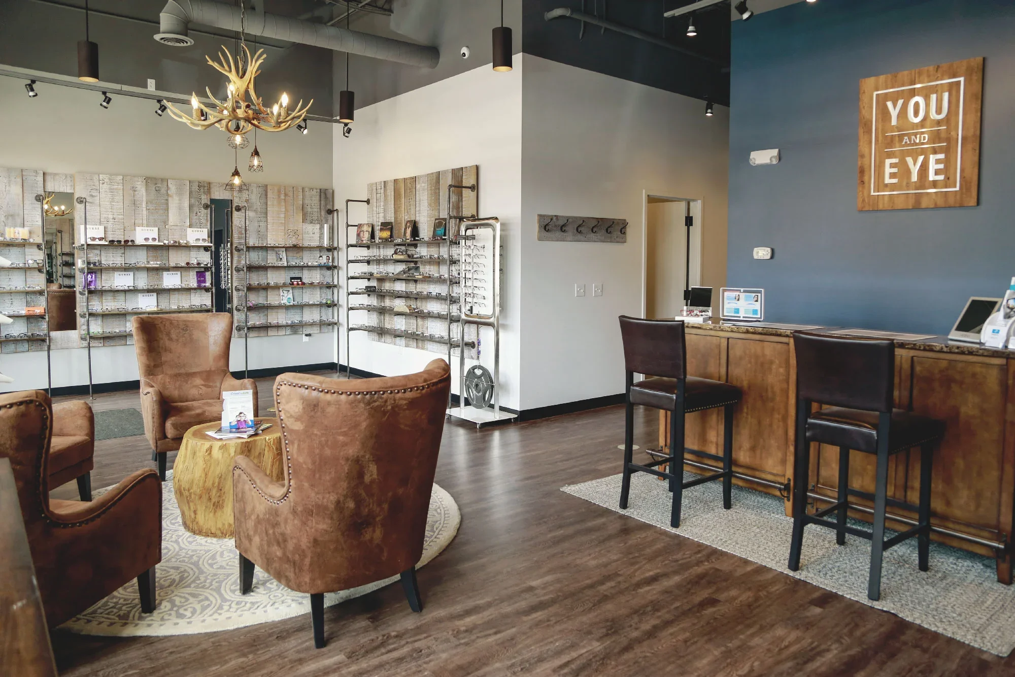 A stylish eyewear store interior with leather chairs, wooden floors, and display shelves filled with glasses. There is a wooden counter with computers and a blue accent wall with a sign reading 'YOU AND EYE' in white letters. Modern decorative lighting includes a chandelier and pendant lights.
