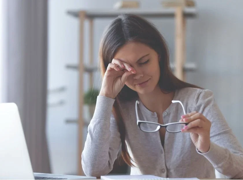 A woman holding her glasses and pressing her forehead, appearing stressed or tired, in an indoor setting with a blurred background.