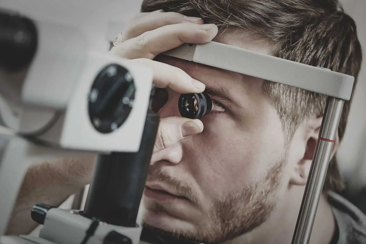 A young man with light brown hair and facial hair using a microscope for an eye examination or research.