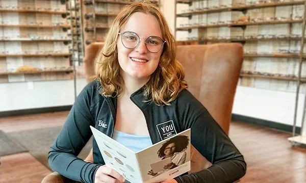 Young woman with glasses sitting in a cozy coffee shop, smiling while holding a brochure, with shelves of coffee bags in the background.