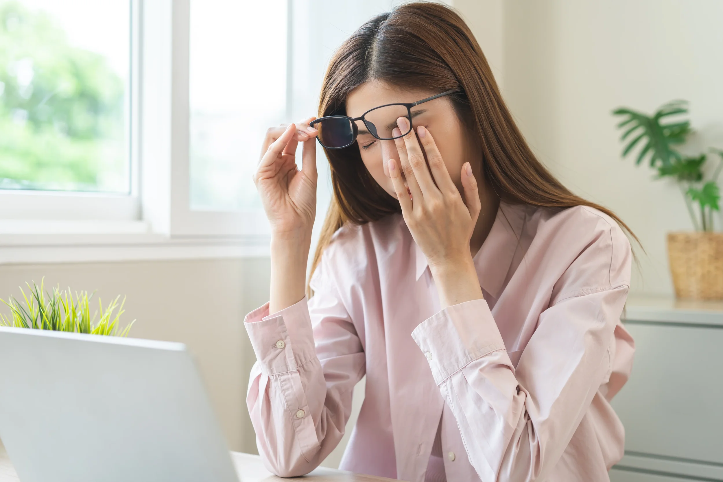 A woman with brown hair wearing a light pink shirt and sunglasses, rubbing her eyes with one hand while sitting at a desk in front of a window with a green plant in the background.
