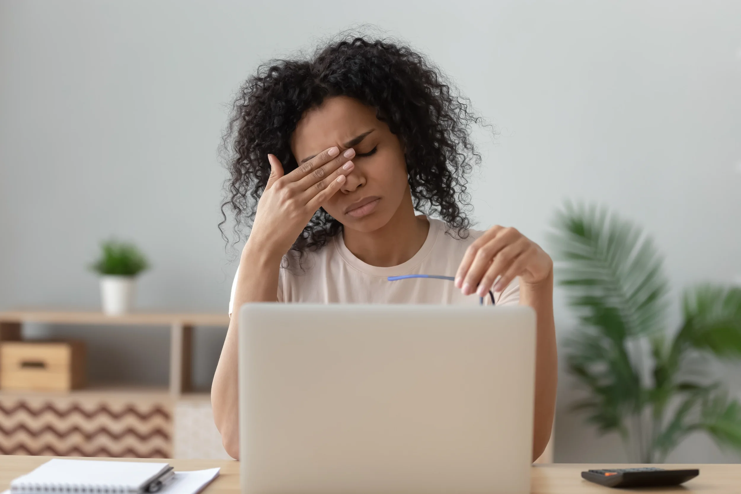 A woman with curly black hair is sitting at a desk, holding her forehead with her left hand in a stressed or tired manner, looking at a laptop in front of her. There are a small notebook and a remote control on the desk, with plants blurred in the background.