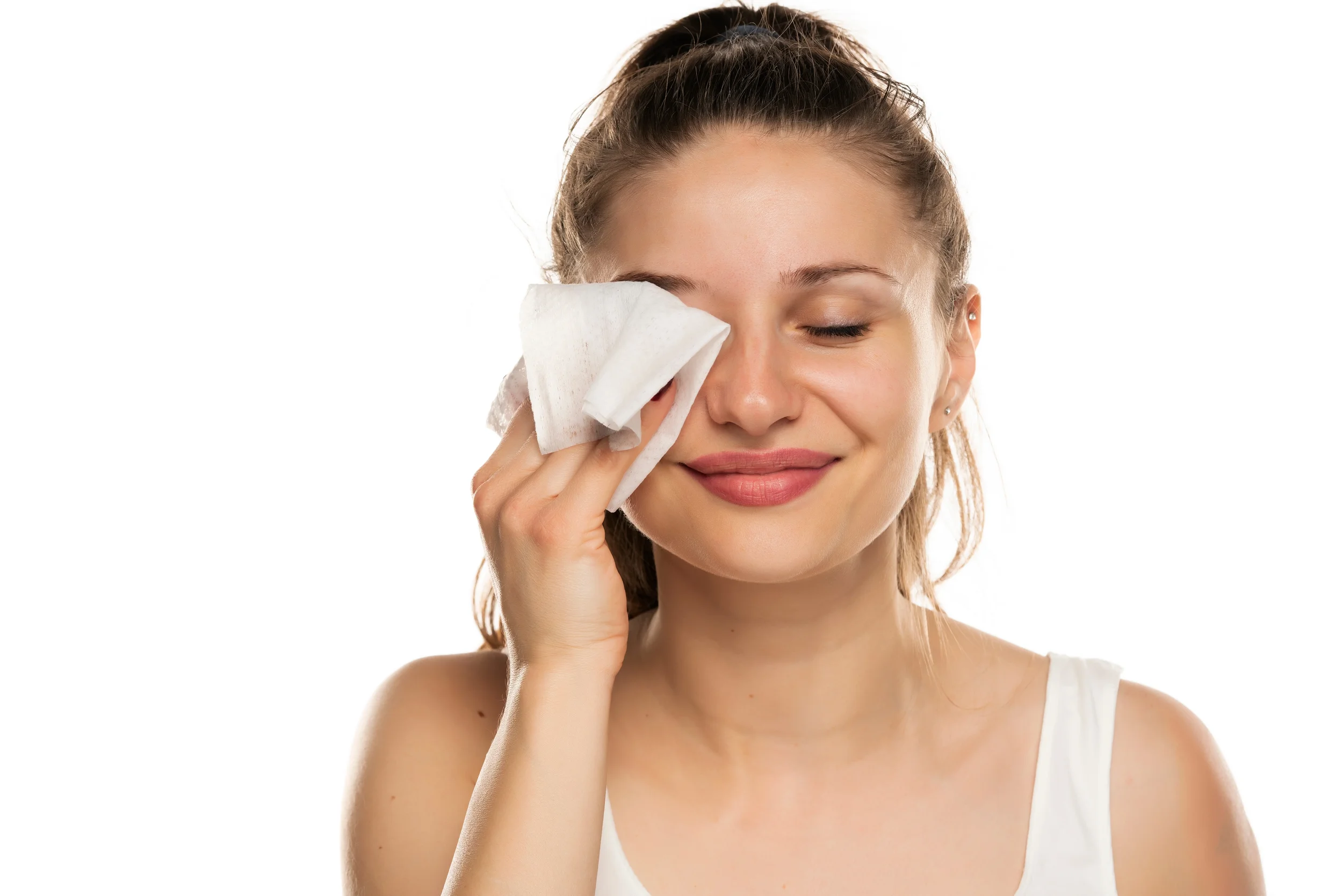 A woman with brown hair in a ponytail is smiling gently with her eyes closed while wiping her face with a tissue or cloth.