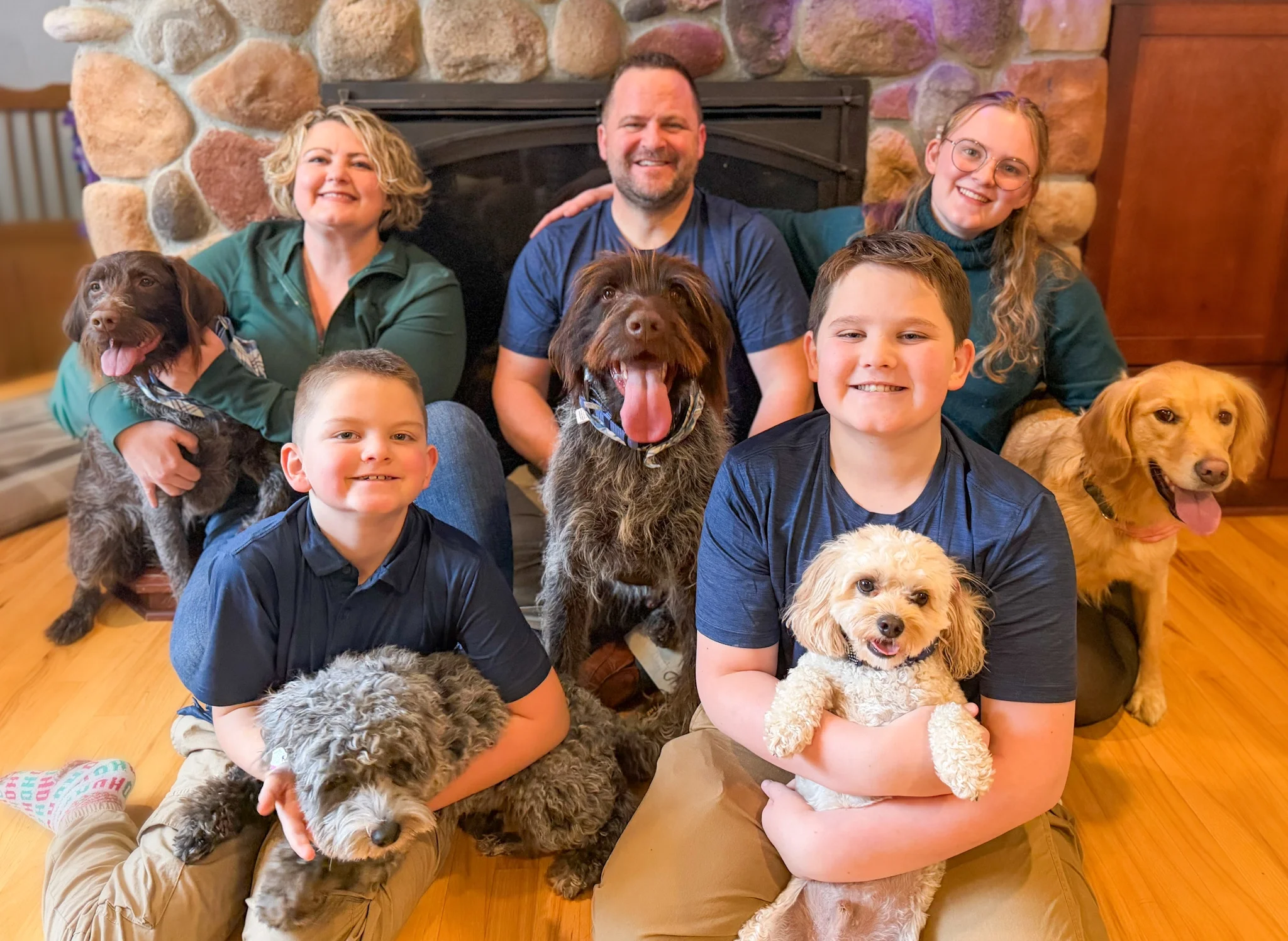A group of five people and eight dogs gathered in a cozy indoor setting with a stone fireplace in the background. The group includes children, adults, and a man with a beard. The dogs are various breeds, mostly brown and fluffy, with one wearing a bandana. Everyone appears happy and relaxed.