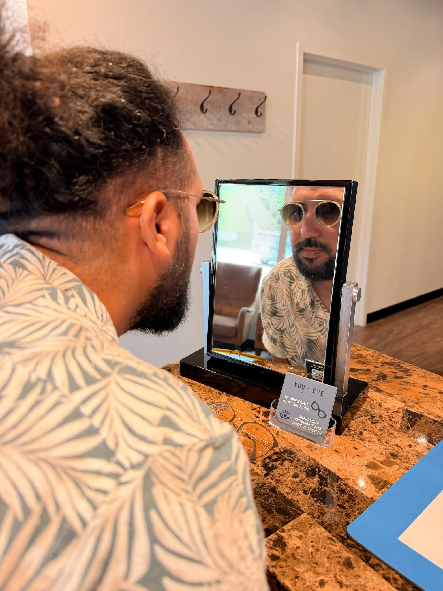 A man with glasses and a patterned shirt is looking into a small mirror at a store. There is a sign on the counter asking for feedback. The background has a white wall with hooks and a door.