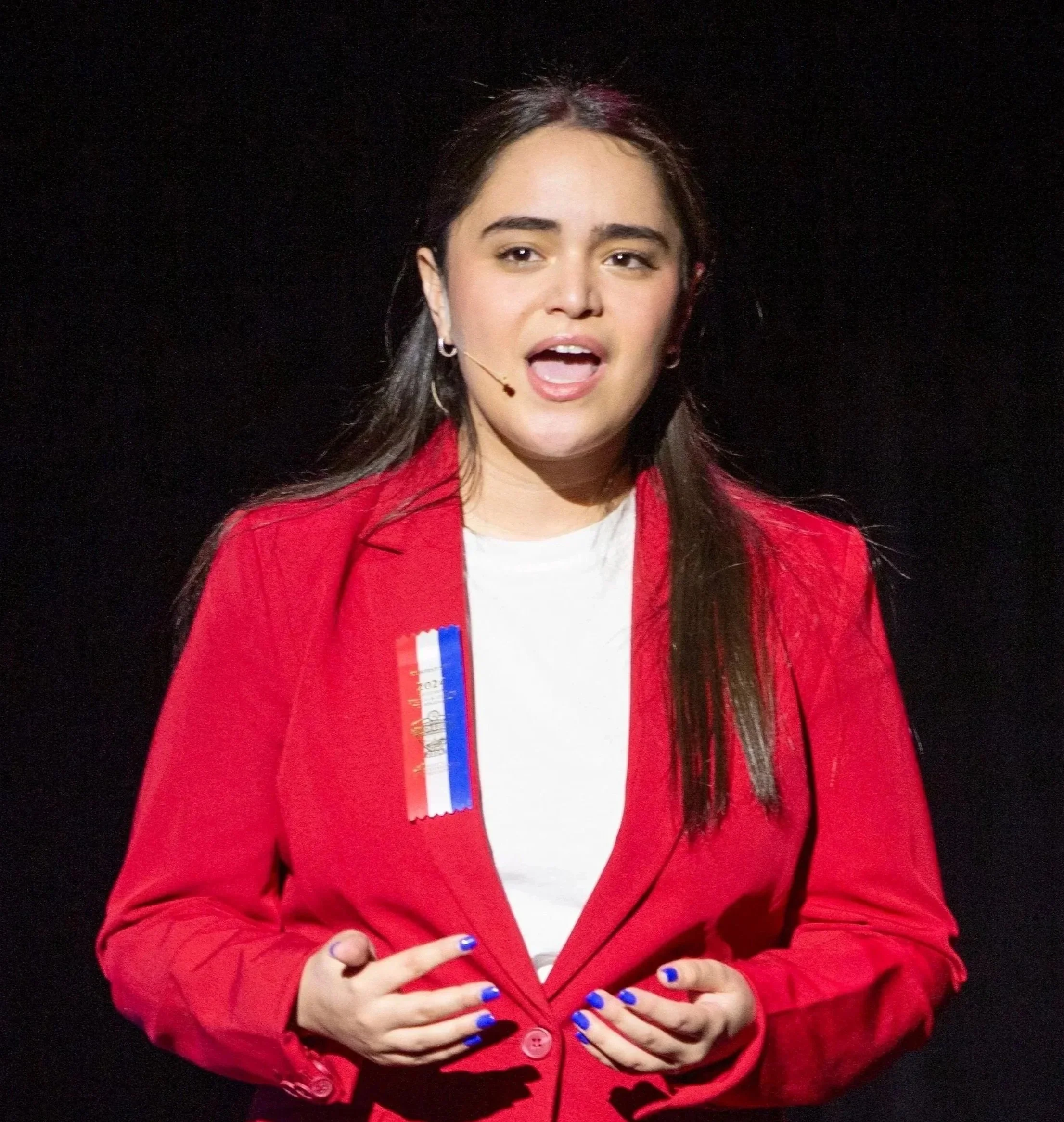 A young woman with dark brown hair and blue nail polish wearing a red blazer and a white shirt, speaking into a headset microphone, with black background.