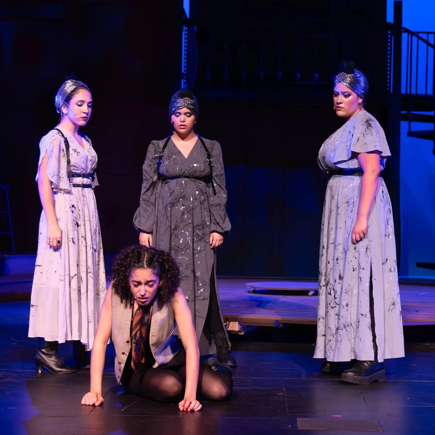 Four women performing on stage in costumes, with one woman kneeling and appearing distressed, surrounded by others with serious expressions under stage lighting.