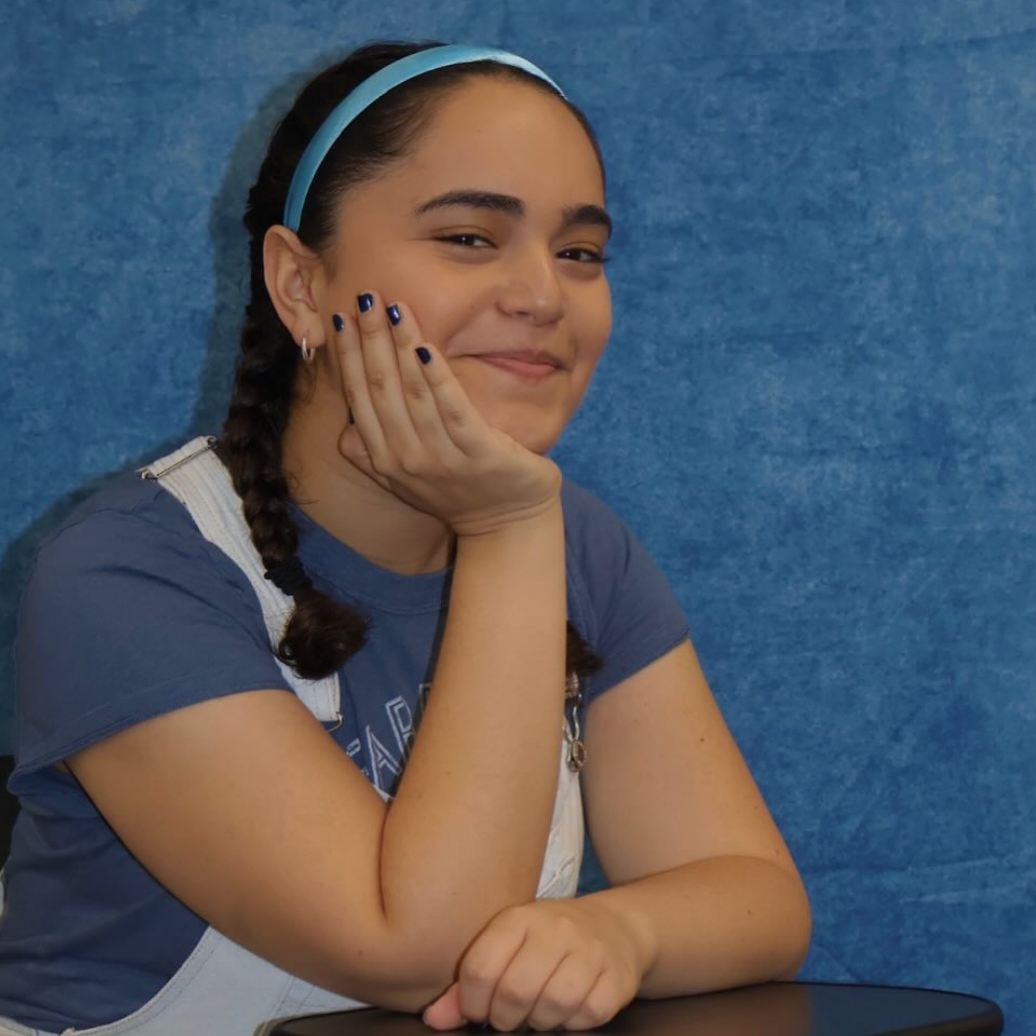 A girl with braided hair, wearing a blue headband and gray shirt, sitting against a blue background, smiling with her hand resting on her cheek.