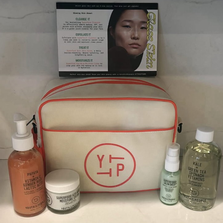 A skincare product box titled Glossier Skin Set featuring a photo of a woman with clear skin. In front, there are various skincare bottles and jars, including a pump bottle labeled Papaya, a small spray bottle, a container labeled Superfood Moisture Cream, a larger bottle of Kale Green Tea Spinach Vitamins, and a tan-colored travel pouch with red trim and a logo that reads