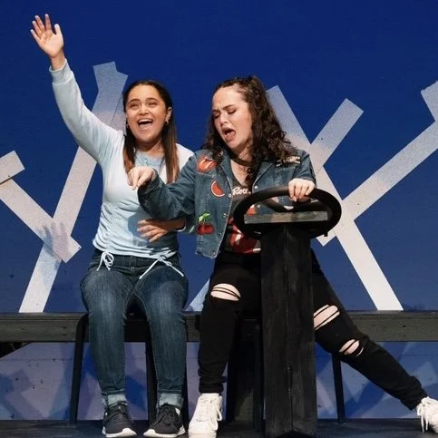 Two young women sitting on a bench, smiling and laughing during a stage performance, with one waving and the other holding the steering wheel of a prop vehicle.