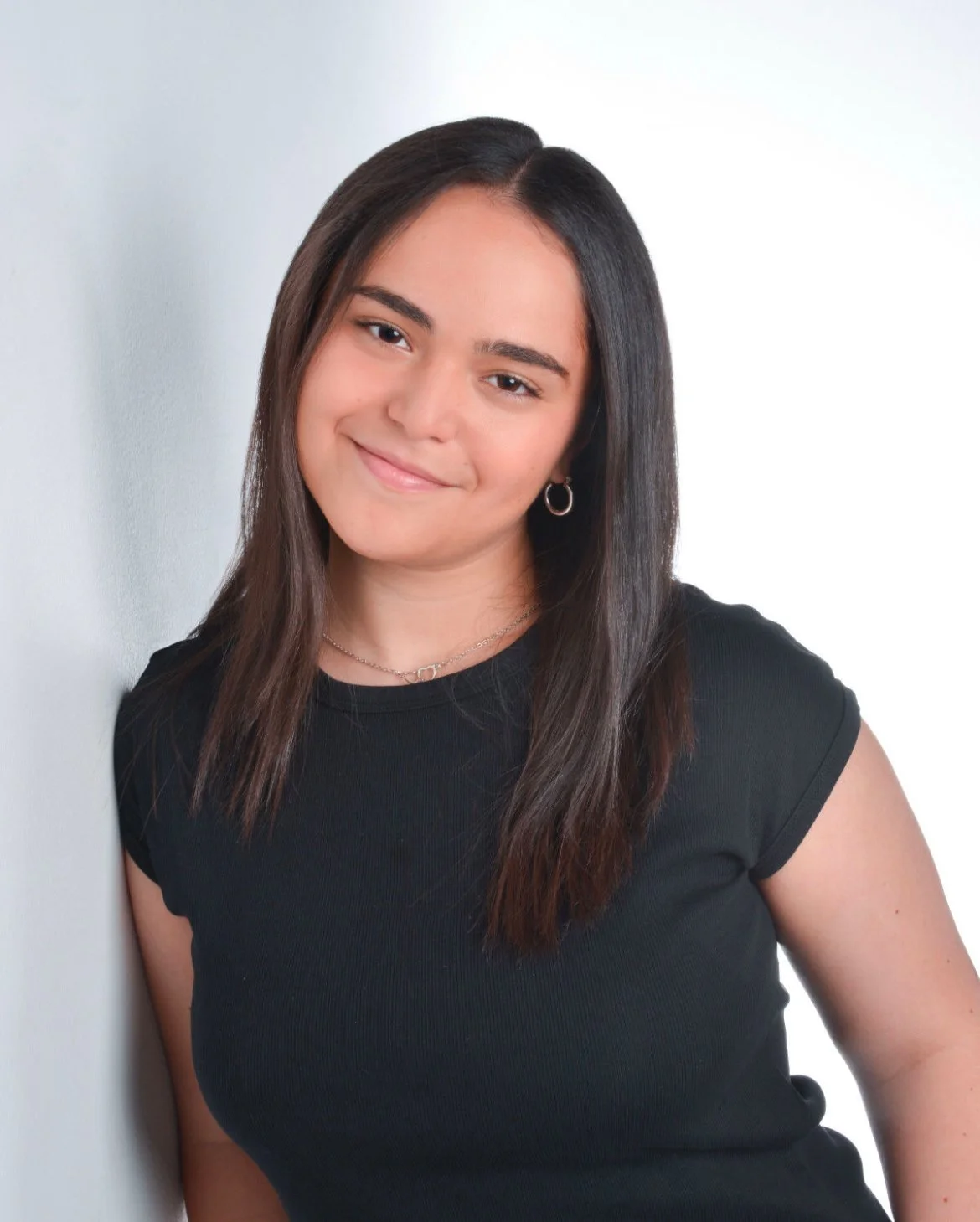 A young woman with long dark hair, wearing a black t-shirt, smiling slightly, standing against a light-colored plain background.