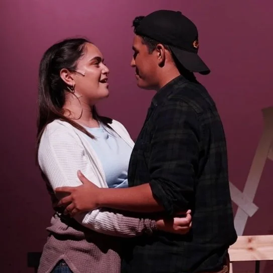 A young woman and a young man are standing close together, smiling, and looking into each other's eyes indoors with a pink background.