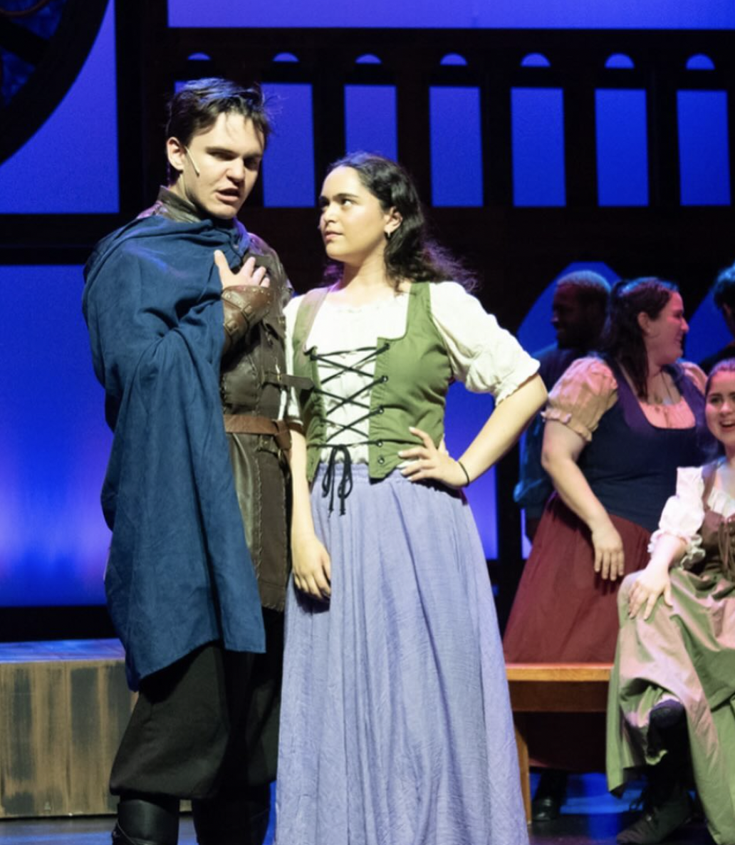 Two actors in costume performing on stage, with several other performers in period costumes in the background.