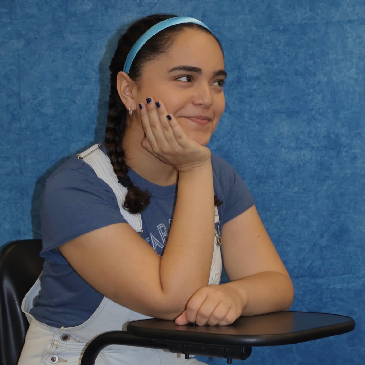 A young woman with braided hair and a light blue headband, sitting with her hand on her cheek, smiling softly, wearing a blue T-shirt and white overalls, against a blue background.