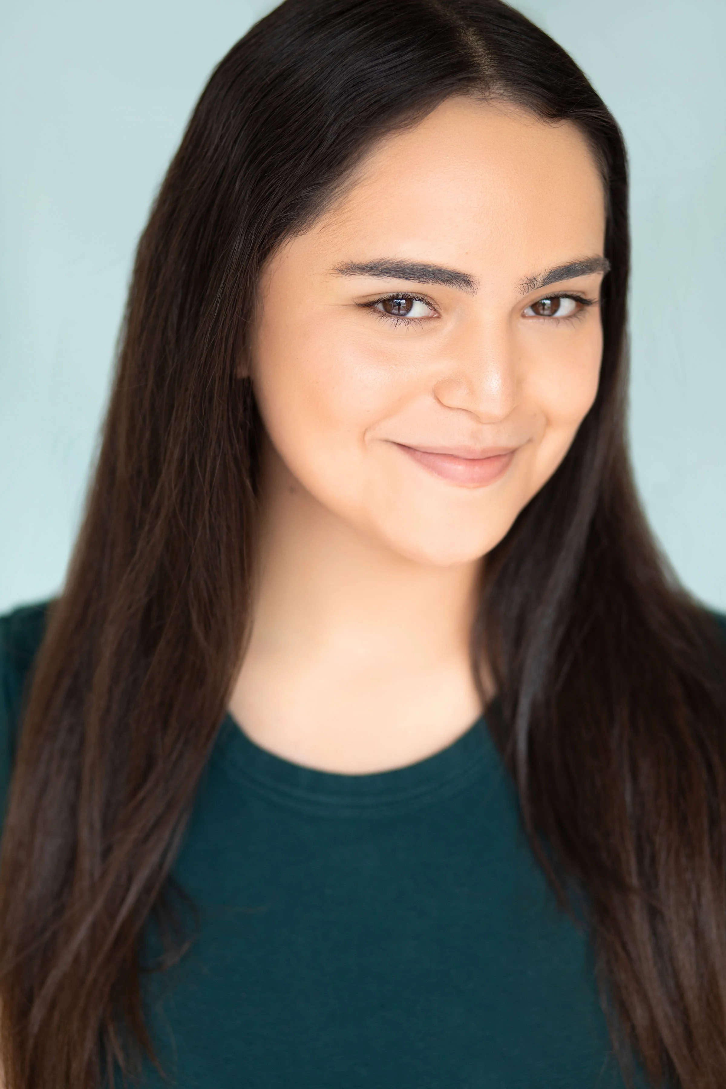 Close-up of a young woman with long dark hair, smiling softly, wearing a dark green top, with a plain light background.