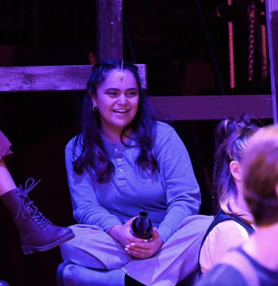 A young woman with long dark hair sitting on the floor, smiling, with purple lighting around her, holding a bottle in her hands.