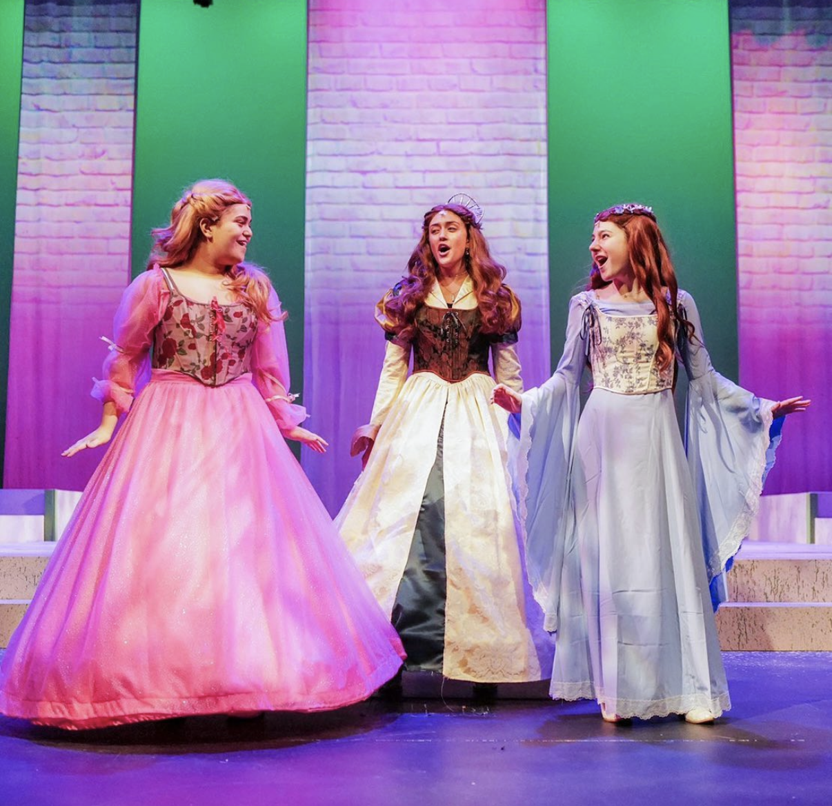 Three women dressed as fairy tale princesses performing on stage, with colorful lighting and a brick wall backdrop.
