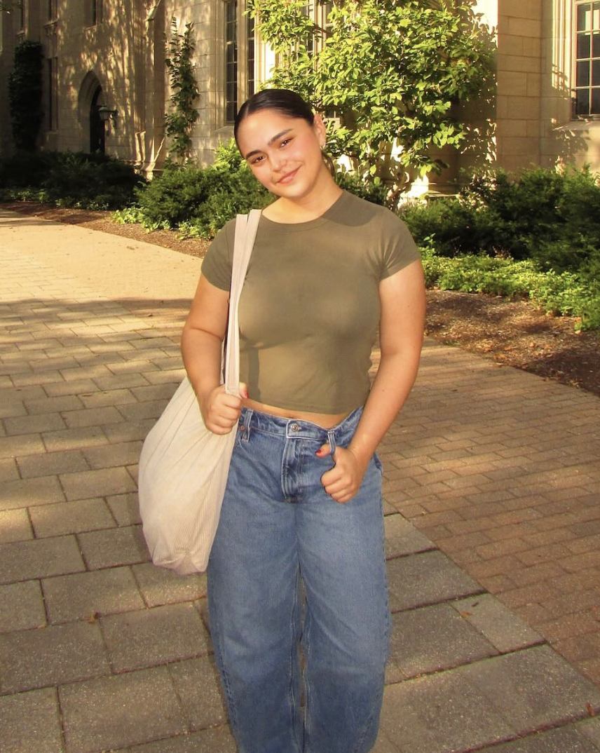 Young woman with dark hair, wearing a green crop top, blue high-waisted jeans, and carrying a light-colored tote bag over her shoulder, standing on a brick sidewalk outside a building with greenery and trees in the background.